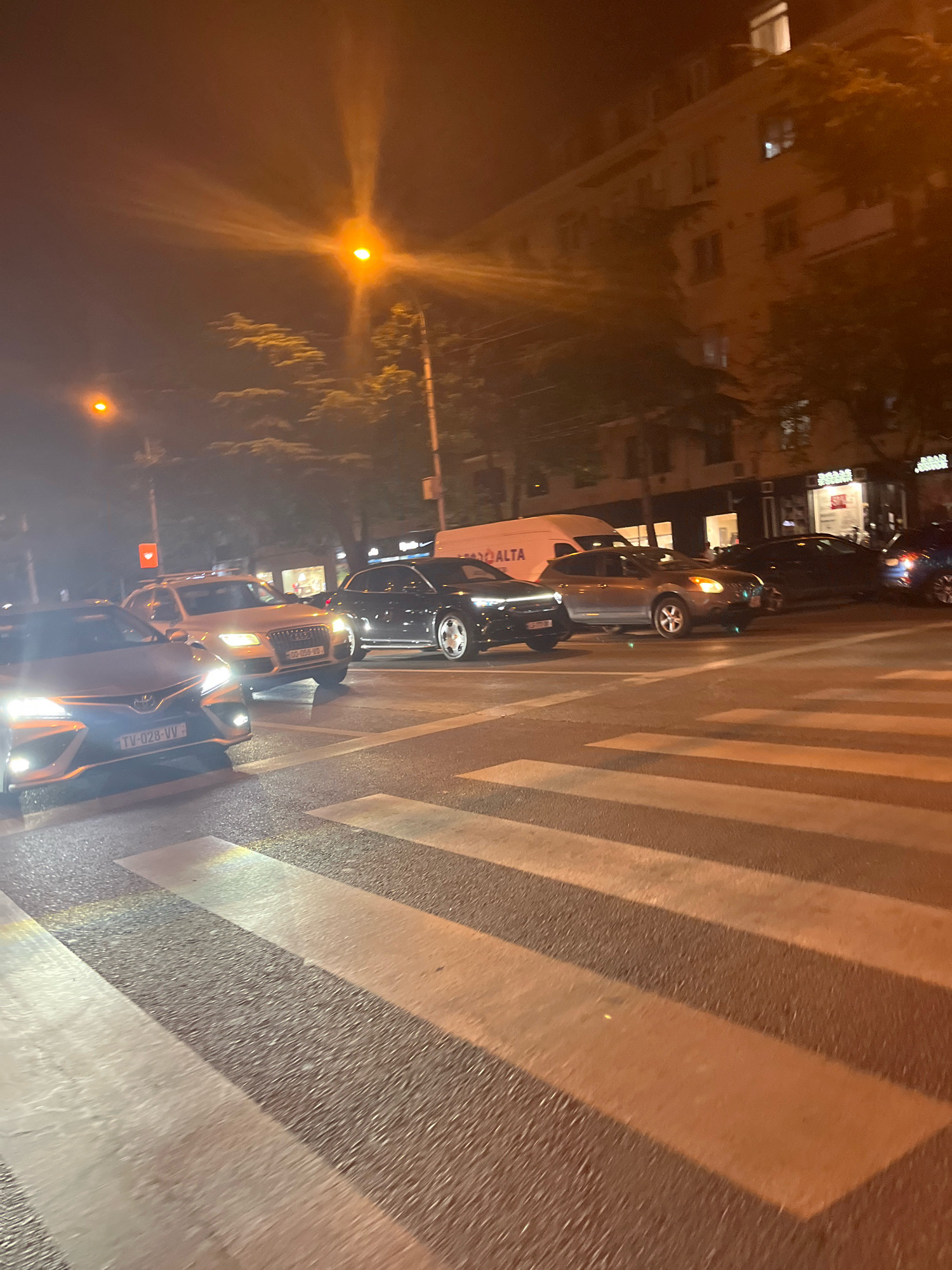 This is a nighttime street scene in Tbilisi, Georgia. The image captures a pedestrian crossing with several cars stopped at what appears to be an intersection. Streetlights cast an orange glow over the scene, creating a hazy atmosphere. The cars visible include a silver Audi SUV, a dark-colored sedan, and a blue car further in the background. In the foreground, the white stripes of the pedestrian crossing are prominent. Buildings and trees line the street, and a white van with "ALTA" written on its side is parked on the right side of the road. The traffic lights are not clearly visible in this frame. The overall impression is one of a typical urban setting at night, with traffic at a standstill.