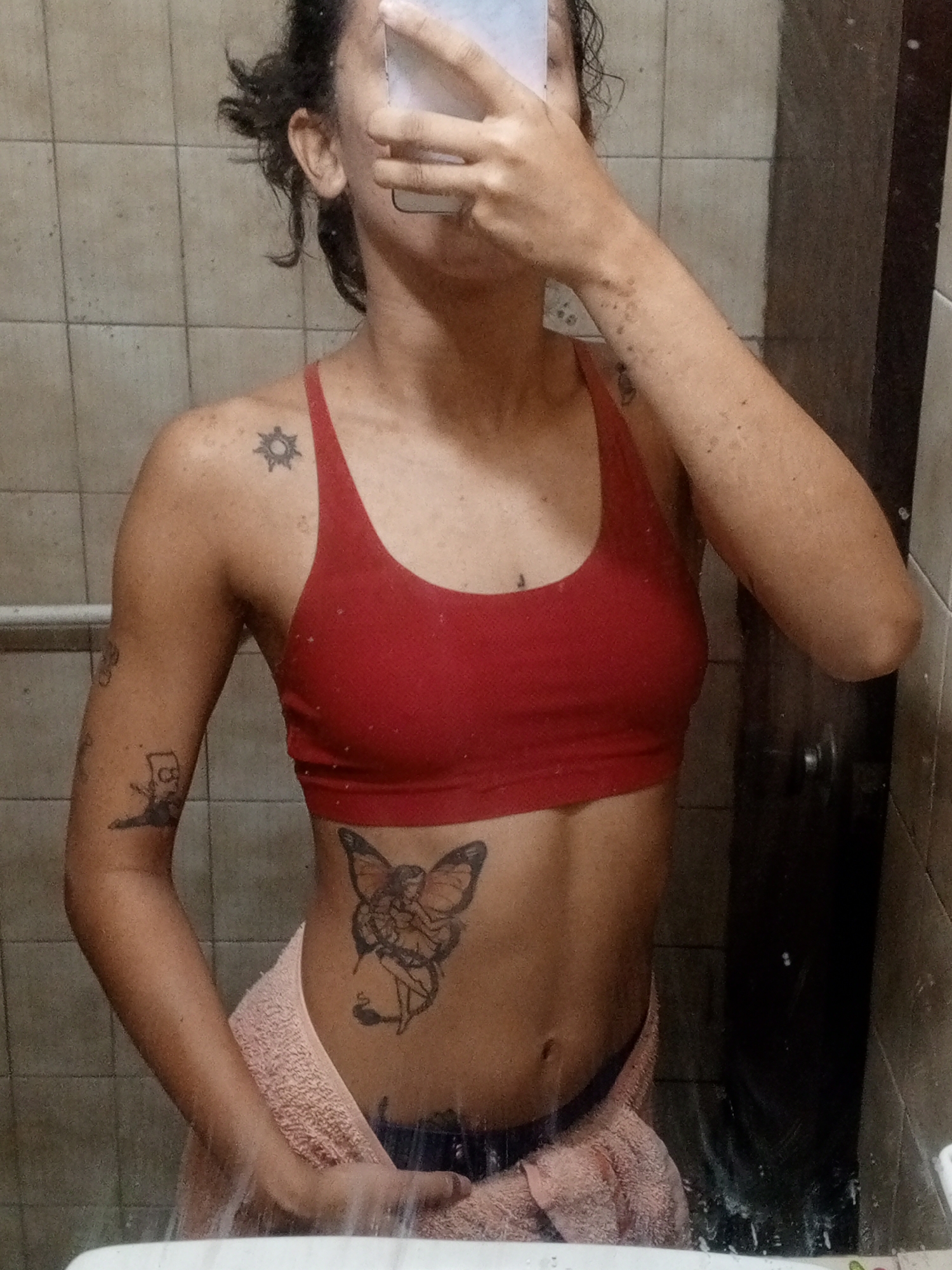 A person stands in front of a mirror, partially obscuring their face with their phone. They are wearing a red sports bra and a towel is draped around their waist. Tattoos are visible on their upper left arm, shoulder, and abdomen. The background features tiled walls and a door. The location in the city of Porlamar, Venezuela cannot be determined from the image.