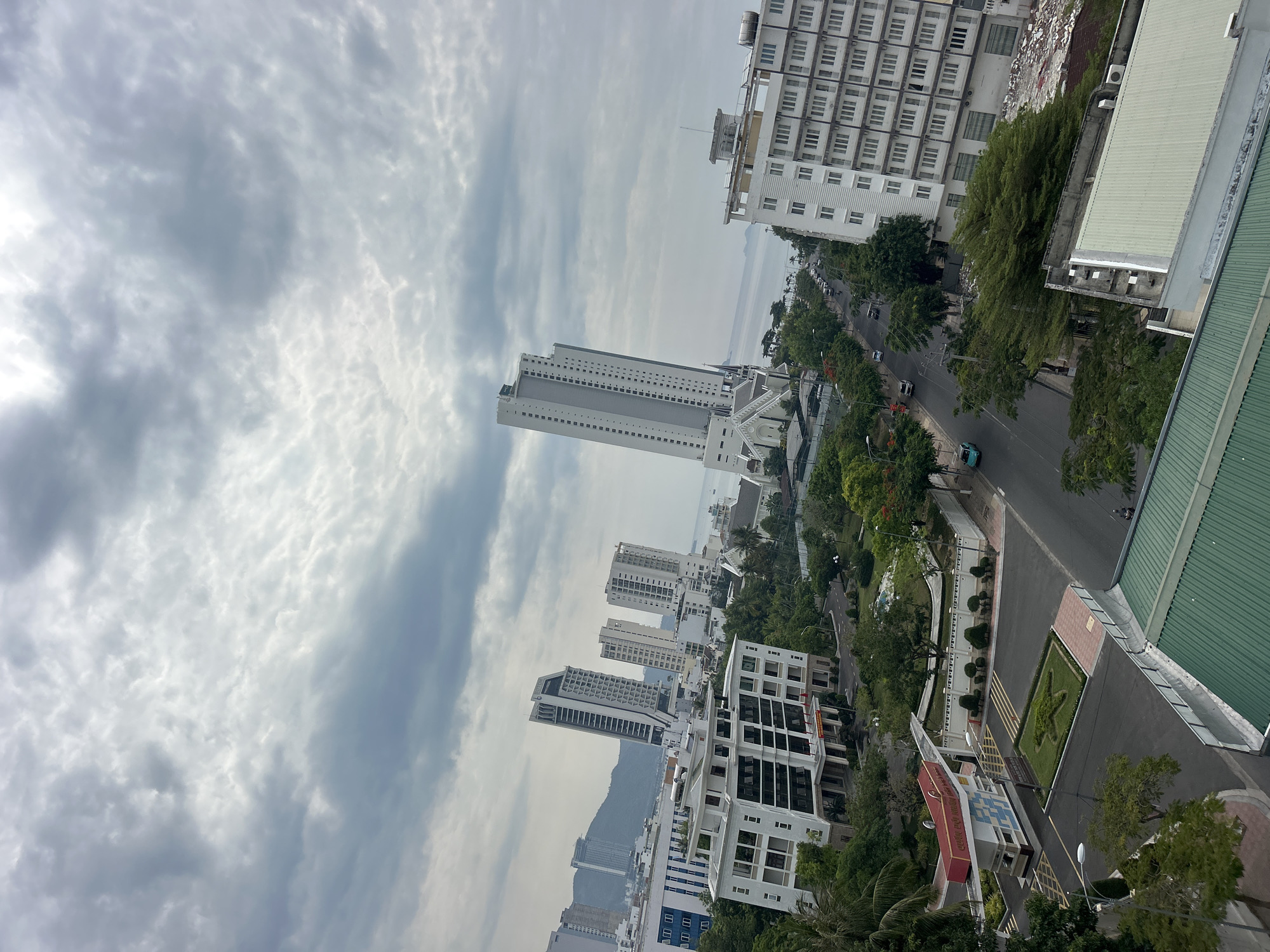 The image presents an elevated view of Bac Nha Trang Ward, Vietnam. The scene is dominated by a street lined with lush trees, running between various buildings. Several multi-story white buildings of diverse architectural styles populate the landscape, ranging from mid-rise to high-rise structures. The sky above is filled with a blend of cloud formations, casting varying levels of light across the urban panorama. A distant mountain range is barely visible in the hazy horizon. Cars are moving down the street. A manicured landscape is featured in the lower right corner. No people can be identified from this height.