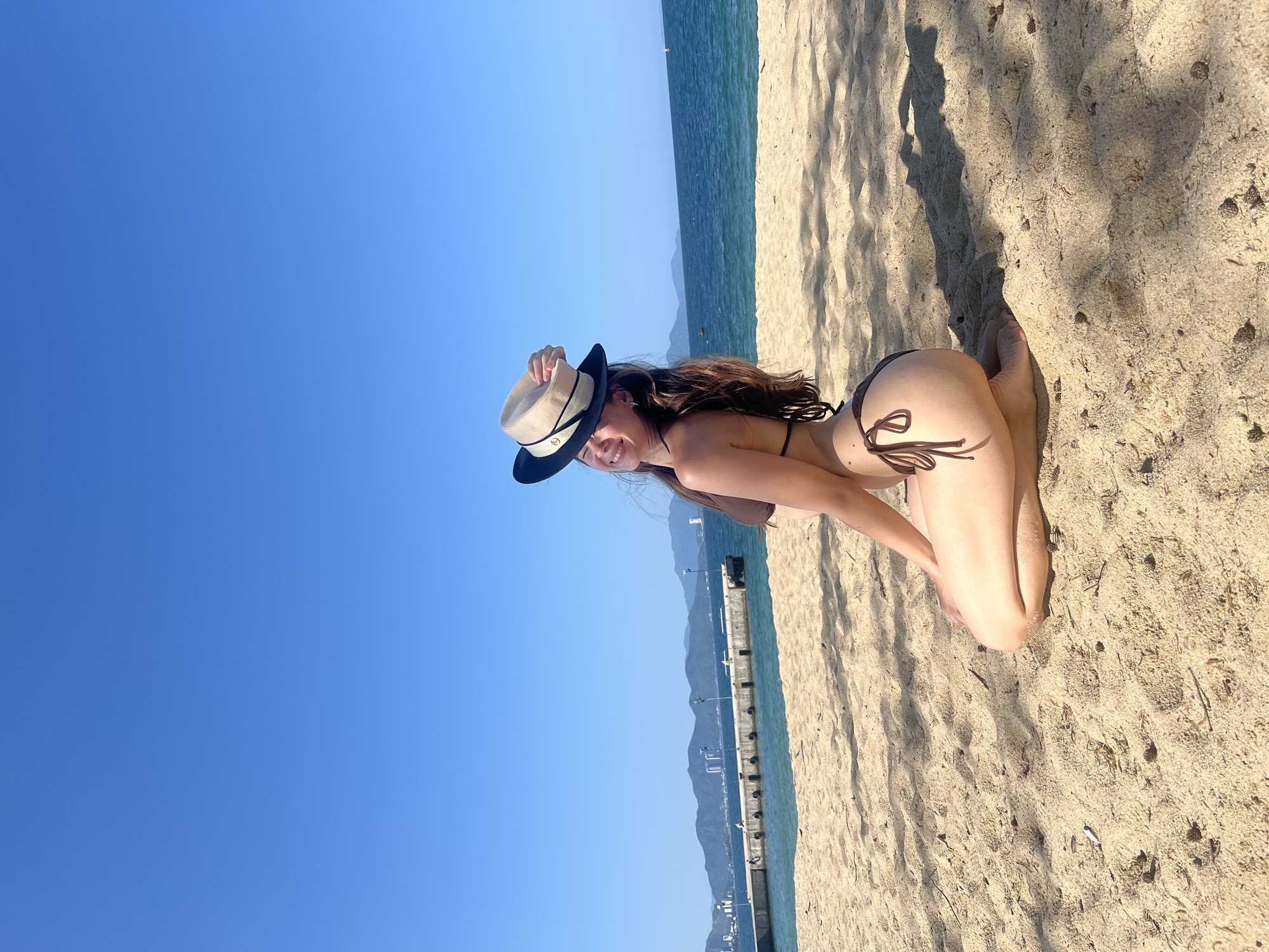A female subject is depicted kneeling on a light brown sandy beach under a clear blue sky in Nha Trang, Vietnam. She wears a brown two-piece bikini and has long brown hair. Her right hand holds a black and beige fedora-style hat positioned above her head. She faces left, with her body angled towards the camera, looking back over her left shoulder with a smile and squinted eyes. Overhanging foliage casts distinct shadows across the sand, which also contains scattered small debris including seed pods and a cigarette butt. In the midground, calm blue ocean water extends towards the horizon. A concrete pier, equipped with several light posts, extends from the left side of the frame into the water, with multiple indistinct human figures visible on its surface. Distant mountain ranges form the backdrop, with urban high-rise structures visible at their base on the left. A small yellow buoy marks a point in the far-right water.