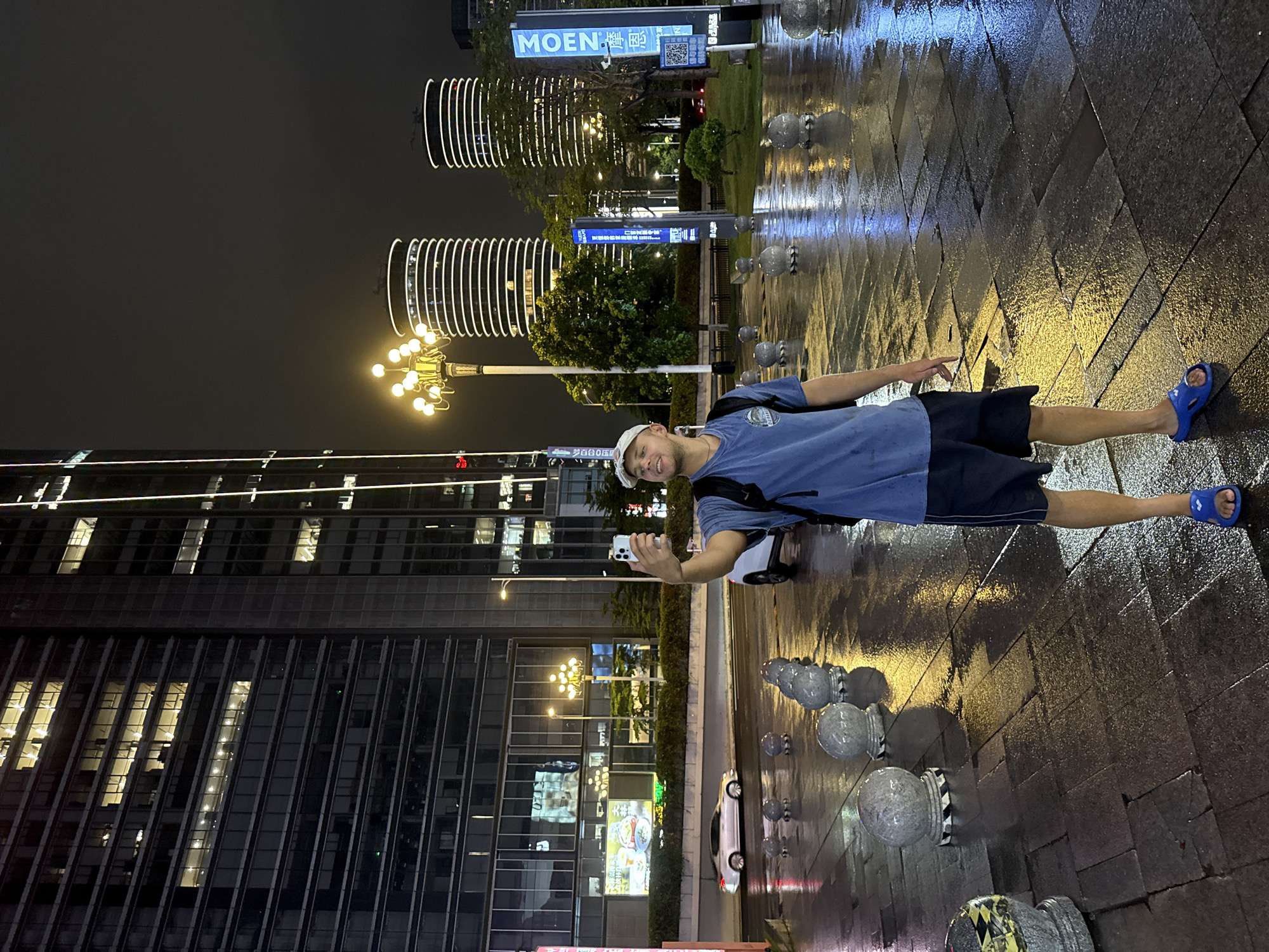 A young Caucasian male stands on a wet, paved city sidewalk at night, taking a selfie with his smartphone. He is wearing a blue t-shirt, dark shorts, a white baseball cap, and blue flip-flops. A black backpack is slung over his right shoulder. The sidewalk is illuminated by streetlights and the reflections of surrounding buildings. Behind him, a multi-lane road is visible with a silver car driving in the distance. In the background, there are tall modern buildings with illuminated windows, and two distinctive cylindrical buildings with white horizontal light strips. A decorative lamppost with multiple lights stands to the left of the cylindrical buildings. The scene appears to be in Tianhe District, China, with signage visible in Chinese characters.