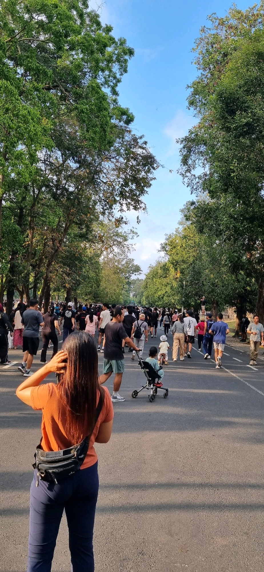 A group of people are walking along a paved road lined with trees, suggesting a park or tree-lined avenue, possibly in Denpasar, Indonesia. The time of day appears to be late afternoon or early morning, with soft sunlight casting shadows. A woman in an orange shirt and blue pants, facing away from the camera, is in the foreground. She has a black fanny pack around her waist. In the middle ground, a man is pushing a stroller with a child inside, and another child is riding a small bicycle. Numerous other people, dressed casually, are walking in both directions along the road, some appearing to be exercising or engaging in recreational activity. The trees are lush and green, with a clear blue sky visible above.