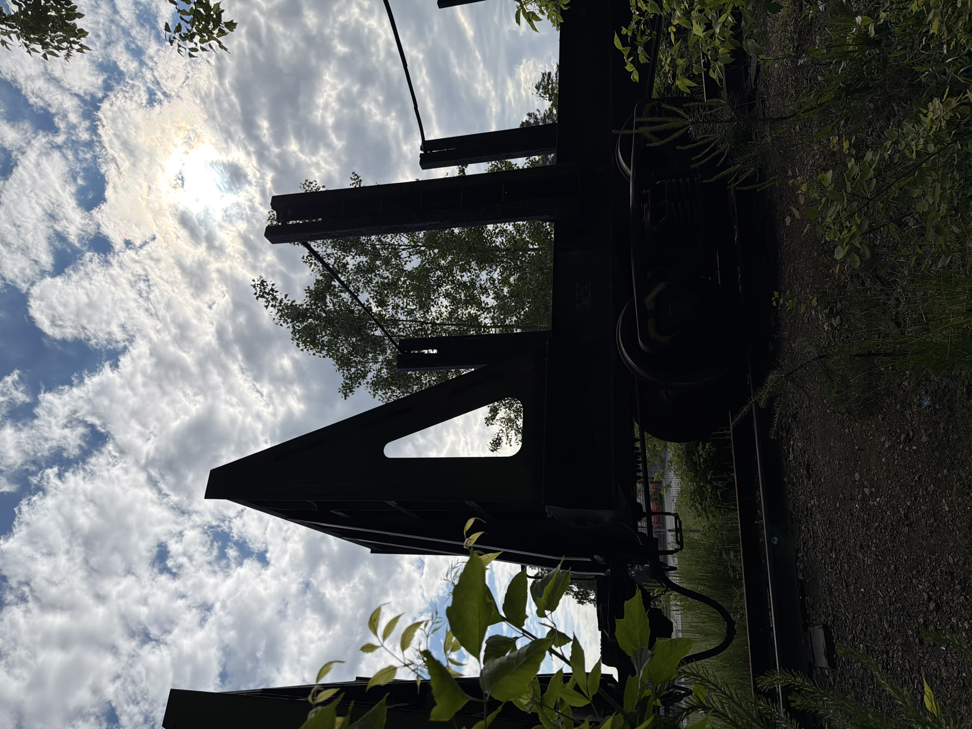 This low-angle photograph captures a large, dark, industrial structure, likely part of an old train car or heavy machinery, silhouetted against a bright, cloudy sky. The image is oriented vertically with the sky at the top and the ground at the bottom.
**Subjects:** The prominent subject is a massive, dark metal framework with distinct lines and sharp angles, occupying the lower half of the frame. On the right side, wheels are visible, confirming its nature as rolling stock. Surrounding the machinery and extending into the foreground and background is abundant green foliage, including tree branches and leaves. Some leaves are close to the camera, appearing slightly blurred, while distant trees are silhouetted against the bright sky. The upper half of the image is dominated by a dynamic sky filled with white and grey cumulus clouds. A brilliant sunburst, mostly obscured but radiating intense light, shines through a break in the clouds slightly to the left of the center.
**Activity or event taking place:** The scene is static, depicting a piece of seemingly abandoned or disused industrial equipment nestled within an overgrown natural environment. There is no human activity.
**Notable details:** The strong sunlight breaking through the clouds indicates it is daytime, likely a bright morning or afternoon. The weather is partly cloudy, creating a dramatic contrast between the illuminated sky and the shadowed machinery. The overgrown vegetation around the heavy equipment suggests a place where nature is gradually reclaiming industrial remnants. The location context of Chaikovsky, Russia, combined with the visible machinery, hints at a former industrial site, railway yard, or perhaps an outdoor display. The low perspective emphasizes the imposing scale of the structure against the vast sky. No discernible text is visible on the machinery.