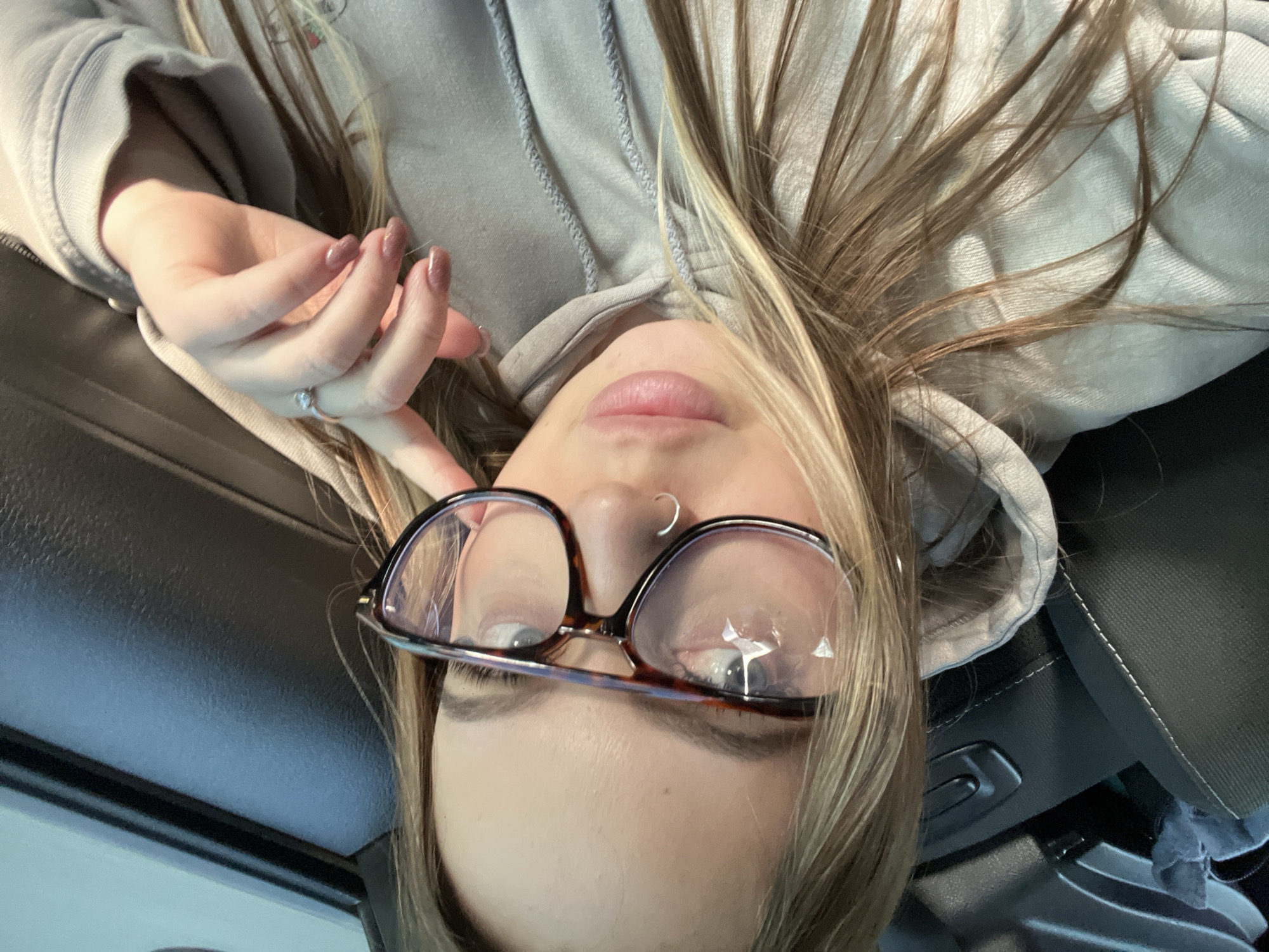 An overhead selfie captures a young person lying down, looking directly into the camera. The subject has long, light brown and blonde hair, styled to fall around their head. They are wearing a light grey hooded sweatshirt. A hand with neatly manicured, shimmery light brown fingernails and a silver-colored ring on the ring finger is visible near their face. They wear large, tortoise-shell framed eyeglasses resting on their nose, and a silver hoop nose piercing is also visible. The person's lips appear softly colored with a hint of gloss. The expression is neutral and relaxed. The background features dark, textured surfaces, likely car upholstery and interior panels with some stitching and controls, suggesting the person is inside a vehicle, possibly lying back in a car seat. Bright sunlight illuminates parts of their hair and hand, indicating daytime. No text is visible in the image. The location context provided for this image is Ternovskaya, Russia.