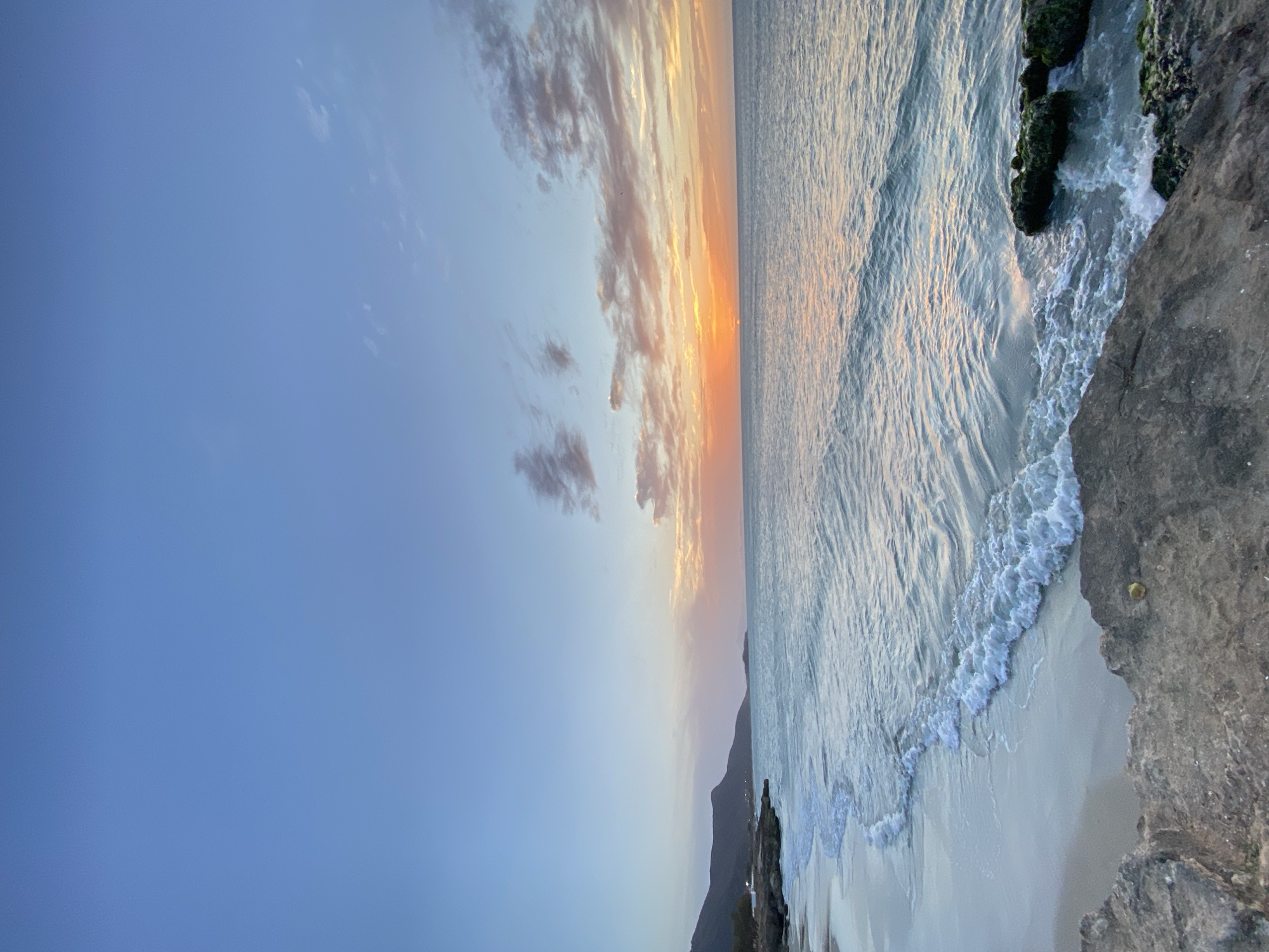 The image shows a coastal scene at sunset in Juan Griego, Venezuela. The sky transitions from blue to orange near the horizon, with scattered clouds. The ocean reflects the sunset colors, and gentle waves break on the sandy beach. Rocks and vegetation are present along the shoreline. A dark hill or mountain can be seen in the background.