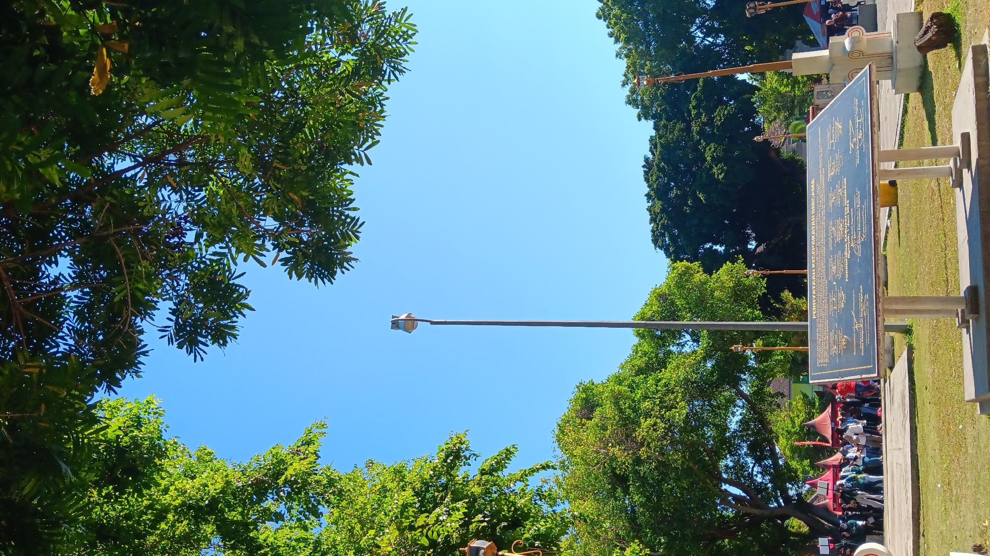 This is a photograph taken outdoors on a bright, sunny day, likely in the city of Nganjuk, Indonesia, given the text on the plaque. The scene is dominated by a clear blue sky, with the upper edges of the frame filled with lush green tree canopies. In the foreground, a large, dark-colored plaque is mounted on two light-colored stone pillars. The plaque has text printed on it, suggesting it's an informational or commemorative display. The text on the plaque appears to be in Indonesian, with headings like "PERNYATAAN KEGIATAN BERSAMA" and "FORKOPIMDA KABUPATEN NGANJUK".
Behind the plaque, a tall, slender pole with a light fixture at the top stands prominently in the center of the image. To the left and right of this central pole, and further in the background, are more trees, some with visible structures that appear to be lamps or decorative poles. In the middle ground, to the left of the central pole and behind the plaque, there's a gathering of people. They are standing near several red and white tents, suggesting some kind of event or market. Some individuals are wearing masks. The ground is covered in green grass, with a paved area visible around the plaque and in front of the tents. The overall impression is of a public space, possibly a park or town square, with a historical or informational marker.