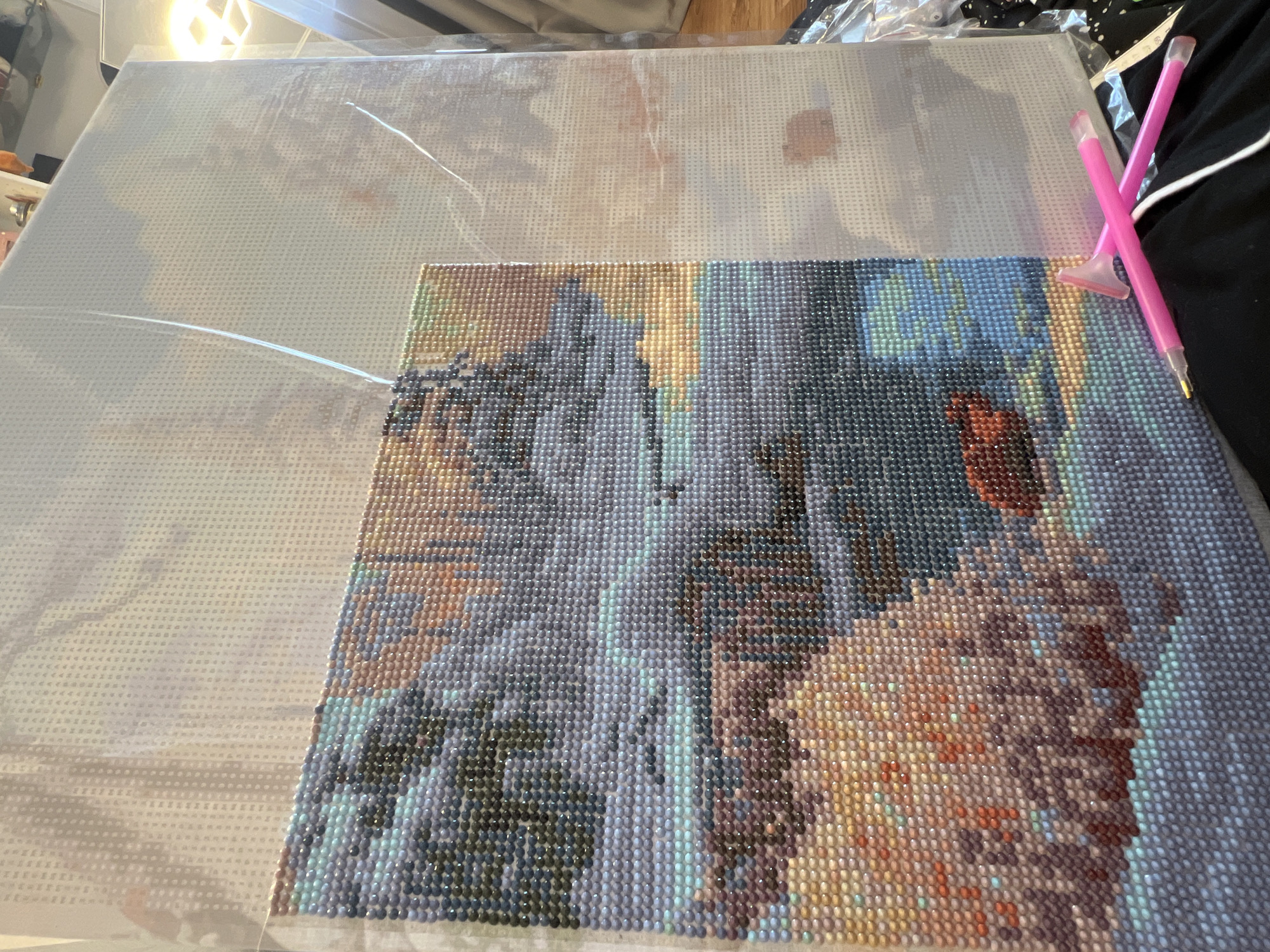 A close-up, high-angle shot captures a diamond painting project in progress, likely in Sochi, Russia. The large rectangular canvas occupies most of the frame, with approximately one-third of the bottom right portion completed with small, faceted resin 'diamonds.' This completed section reveals a vibrant and detailed image, possibly an abstract landscape or a stylized natural scene, rendered in a mosaic of blues, greens, purples, oranges, yellows, and browns. The individual beads shimmer, indicating their reflective nature and creating a textured surface.
The remaining two-thirds of the canvas, towards the top and left, remain uncompleted. This area is covered by a translucent plastic film, protecting the adhesive surface underneath. Through the film, a clear grid pattern is visible, along with faint outlines and symbols that guide the placement of the diamonds.
Two bright pink diamond painting pens or applicators, each with a multi-placer tip, rest on the right edge of the completed section, indicating the tools used for this craft. A bright light fixture is reflected in the top left corner, illuminating the canvas. The background consists of dark fabric and some plastic packaging, suggesting an indoor setting where the crafting is taking place. The scene conveys a sense of quiet, focused activity.