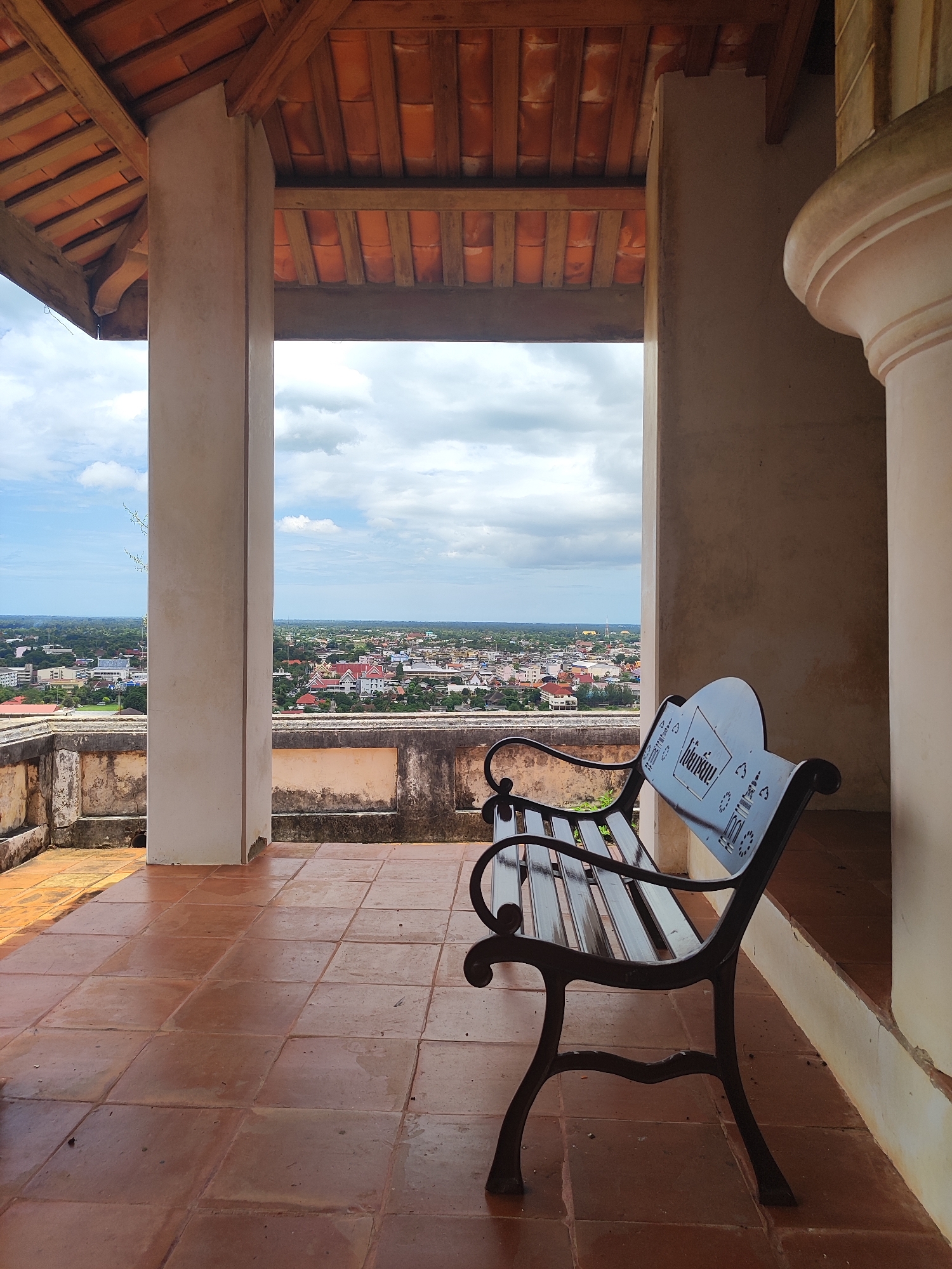 The image shows a shaded outdoor seating area with a view of the city of Phetchaburi, Thailand. In the foreground, a black metal bench with intricate scrollwork on its legs and backrest sits on a terracotta tiled floor. The bench has a heart-shaped back with decorative cutouts and a Thai inscription. To the right of the bench, a large, curved, beige column is visible. The seating area is covered by a roof with wooden beams and orange tiles. Through an opening framed by two pillars, a panoramic view of Phetchaburi unfolds. The cityscape features numerous buildings with red and white roofs, nestled amidst lush greenery. In the distance, the landscape stretches towards a hazy horizon under a partly cloudy sky. The lower portion of the framing structure features a weathered stone balustrade. There are no people present in the image.