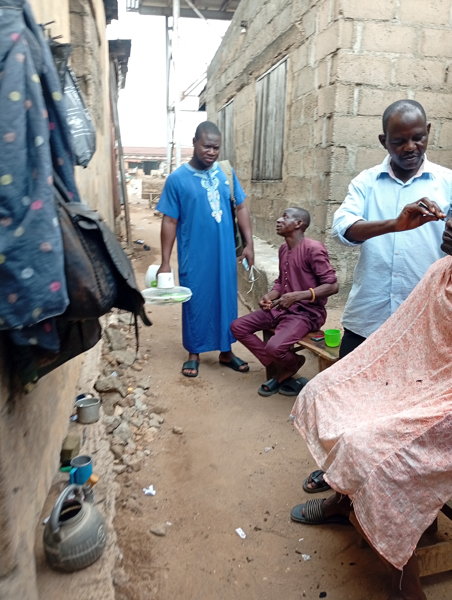 This image captures a vibrant outdoor scene in a narrow dirt alleyway, characteristic of a communal area in Zango-ndaji, Nigeria. The bright, natural daylight indicates it is daytime.
The central activity appears to be a street barbering session. On the right, a man in a light blue checkered shirt and dark trousers, presumably the barber, stands focused on a client. The client is seated, largely obscured by a pinkish-peach patterned cloth, which serves as a barber's cape. The barber holds a small, sharp metal tool, possibly tweezers or a fine clipper, suggesting precision work.
To the left of the barber, another man, dressed in a maroon traditional top and matching trousers, is seated on a low wooden bench. He has distinctive white marks on his face, perhaps a grooming residue or traditional markings, and is looking upwards and to the left. A green plastic cup is visible on the ground next to him.
Further to the left, a third man stands, clad in a flowing blue traditional garment adorned with white embroidery. He holds a white object, possibly a small portable fan or a sieve, in his left hand and observes the scene. All three men are wearing sandals.
The setting includes rough, unpaved ground with scattered small stones and debris. Along the left wall, a blue jacket with polka dots and a dark bag are hanging. Below them, near the base of the wall, an old grey kettle, a blue mug, and a small grey bucket are visible. The buildings flanking the alley are constructed from concrete blocks. No discernible text is visible in the image.
