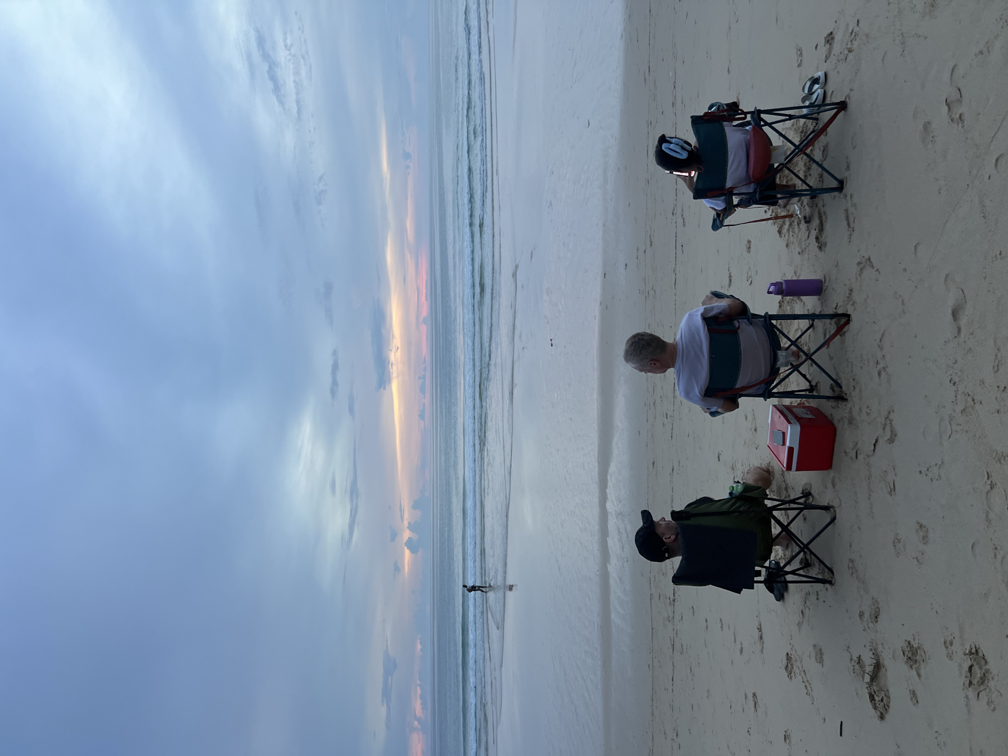 Three individuals are seated in portable chairs on a sandy beach, facing the ocean at dusk. A person in a black baseball cap is seated on the left, facing away from the camera. In the center, an individual with gray hair is seated, also facing away. On the right, a person wearing headphones is seated. A red cooler is positioned between the left and center individuals. A purple water bottle is next to the center individual. A person is walking in the ocean in the background, silhouetted against the horizon. The sky exhibits soft hues of pink and blue, indicating the presence of a sunset. The ocean has gentle waves, and the sand is marked with footprints. This scene is located on the beach in Choeng Thale, Thailand.