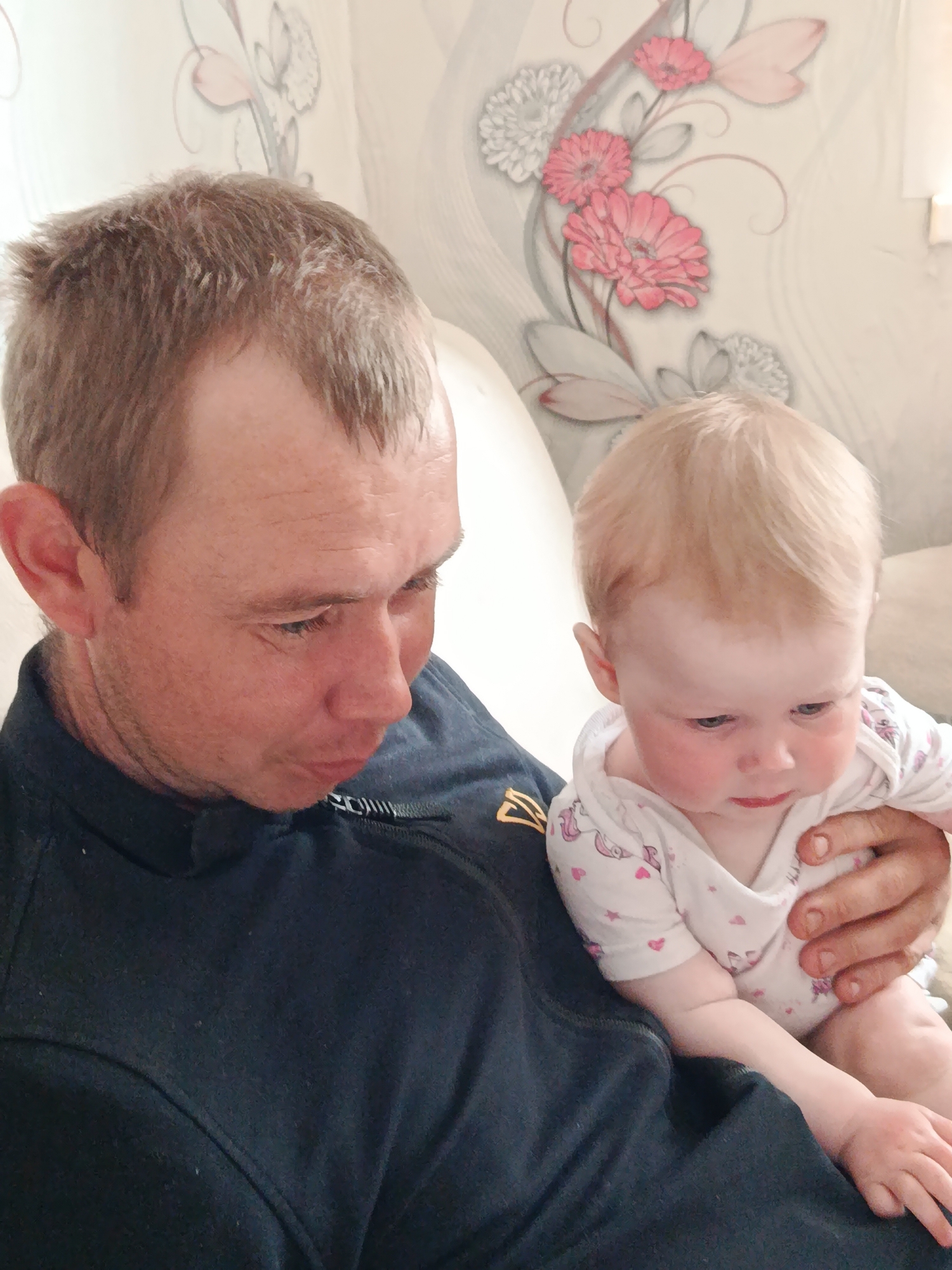 A close-up shot depicts an adult male holding a baby. The man has short, light brown hair and is wearing a dark blue zip-up jacket. The baby, dressed in a white onesie with pink hearts, is resting in the man's arms and looking downwards. The background features a wall decorated with floral patterns. The location is unspecified, Фоки, Russia, is not visible in the picture.