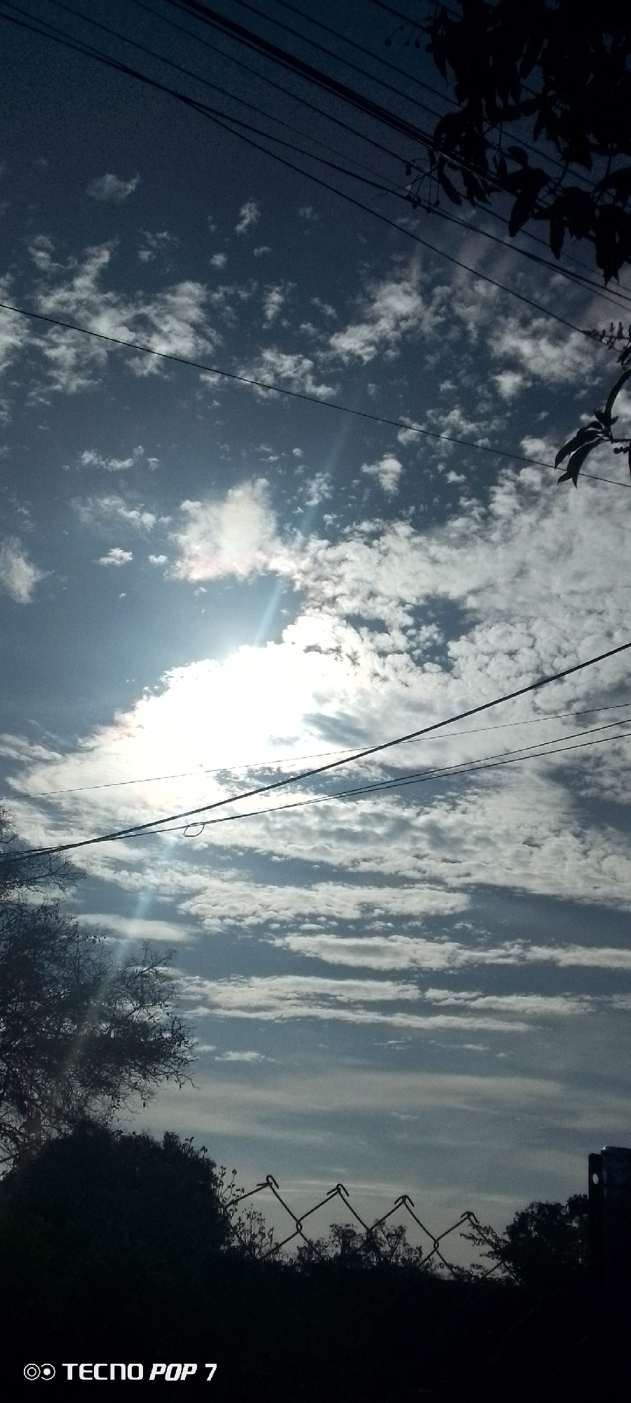 The image depicts a daytime scene in Altagracia, Venezuela. The sky is filled with altocumulus and cirrus clouds, with the sun obscured by cloud cover. Power lines are visible crossing the sky. Below the horizon line, vegetation and a chain-link fence are silhouetted in the foreground. The words "Tecno Pop 7" are displayed in the lower left corner of the image, indicating the device used to capture the photograph. No people are visible.