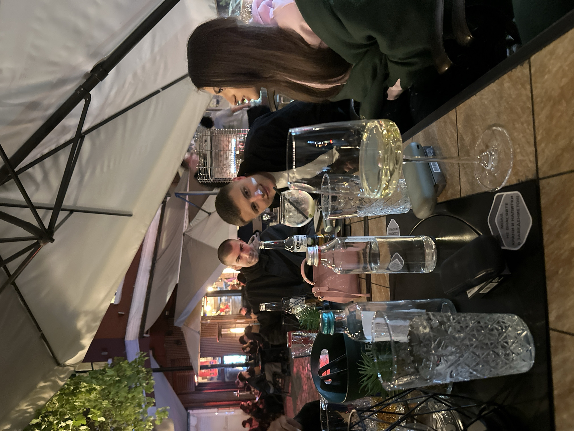 The image depicts a scene at an outdoor restaurant in Plovdiv, Bulgaria. Three individuals are visible at a table under a white parasol. A woman with long brown hair and wearing a green hoodie is seen at the top of the frame. Two men are sitting opposite to her; one is slightly out of focus, while the other, closer to the camera, is wearing a black jacket. The table is filled with glassware including a wine glass with yellow liquid, several bottles (one of which appears to be a Corona Extra), and decorative containers holding plants. The background shows a bustling street scene with people walking, a building with red exterior, and other outdoor dining areas.