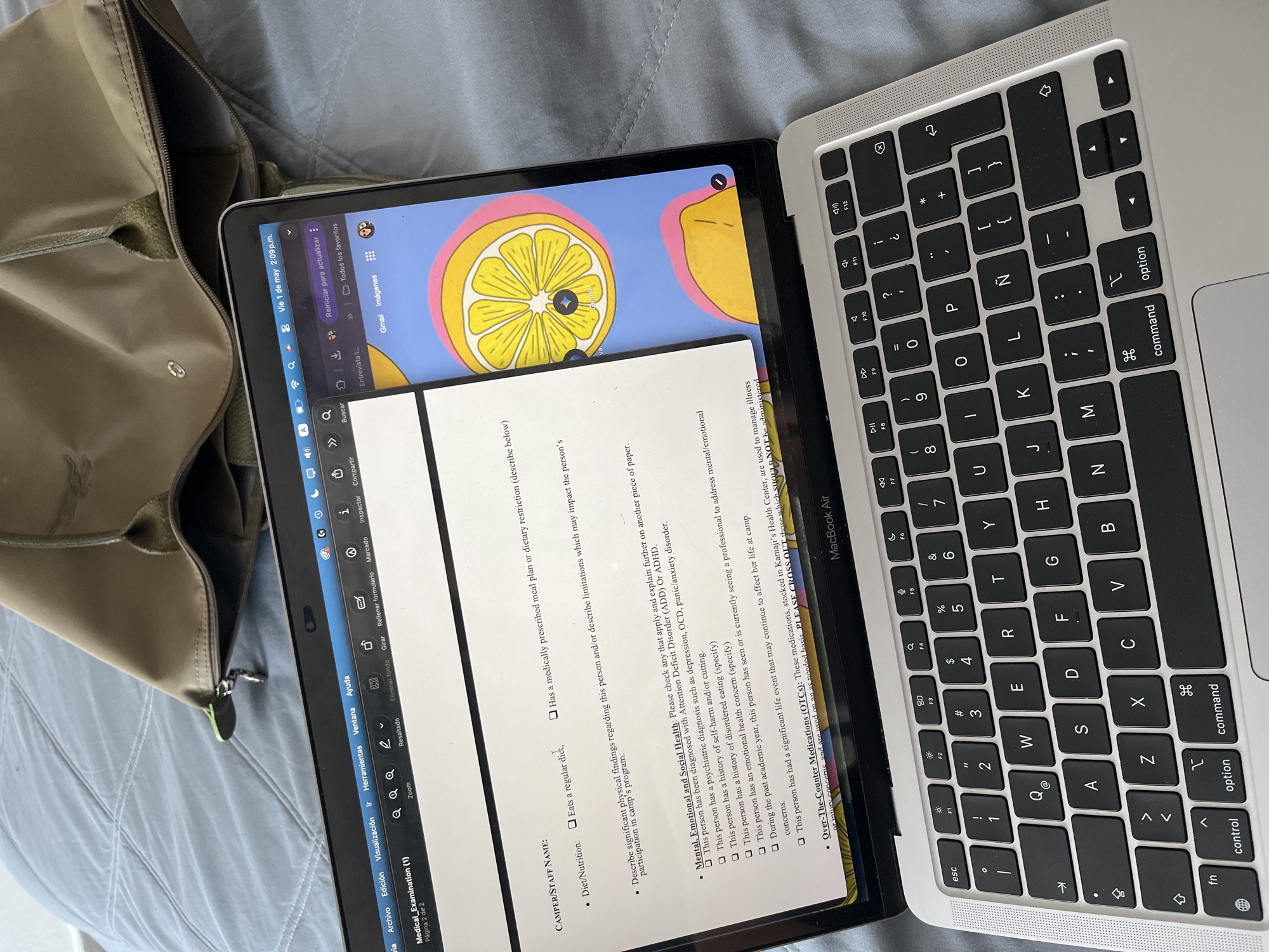 An overhead shot shows an open MacBook Air laptop resting on a light blue/grey quilted surface, likely a bed or sofa. To the left of the laptop, a greenish-brown bag with a zipper is partially visible.
The laptop screen displays a digital document titled "Medical_Examination (1)," which appears to be a health screening form, specifically "Página 2 de 2." The form includes sections for "CAMPER/STAFF NAME," "Diet/Nutrition," and "Mental, Emotional and Social Health." Under "Diet/Nutrition," there are checkboxes for "Eats a regular diet," and "Has a medically prescribed meal plan or dietary restriction." The "Mental, Emotional and Social Health" section lists various checkboxes for conditions like "Attention Deficit Disorder (ADD) Or ADHD," "psychiatric diagnosis such as depression, OCD, panic/anxiety disorder," "history of self-harm and/or cutting," "history of disordered eating," "emotional health concern," and inquiries about professional help or significant life events. Another section is visible for "Over-The-Counter Medications (OTCs)" from "Kamaji's Health Center."
Above the document, a vibrant digital wallpaper featuring a stylized lemon slice on a pink and blue background is visible. The top bar of the laptop screen shows "Vie 1 de may 2:09 p.m.," indicating Friday, May 1st, at 2:09 PM. Other visible elements on the screen's interface include "Reiniciar para actualizar," "Todos los favoritos," "Gmail," "Imágenes," and menu options like "Archivo," "Edición," "Visualización," "Herramientas," "Ventana," and "Ayuda" in Spanish. The MacBook Air's keyboard is visible, featuring a Spanish layout with an "Ñ" key. The setting suggests a casual work or study environment in Puebla, Mexico.