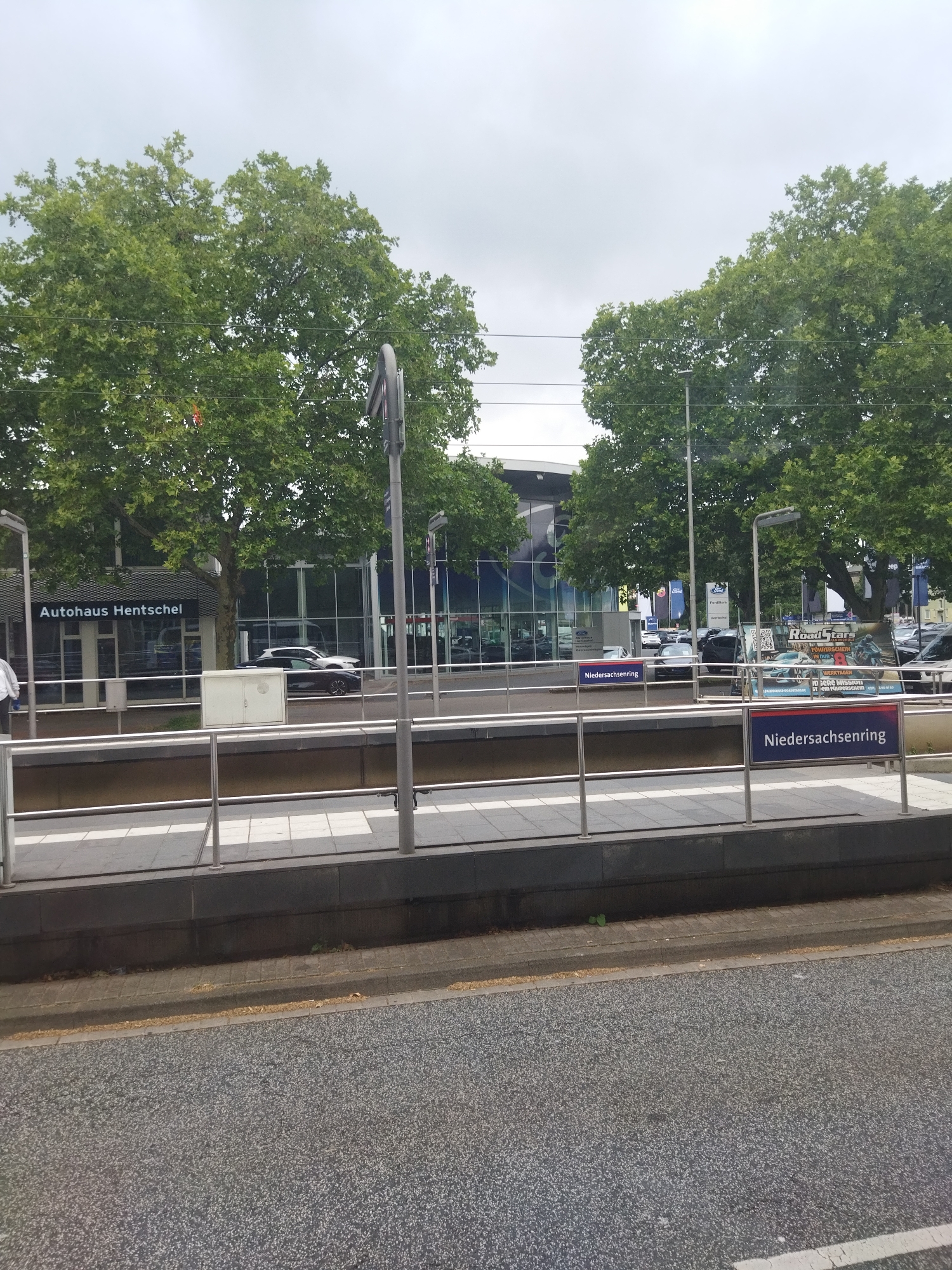 The image shows a street-level view of a public transportation stop in Hanover, Germany, with a focus on signage and surrounding buildings. A blue sign with white lettering reads "Niedersachsenring," indicating the location. To the left, a building facade with large glass windows displays the text "Autohaus Hentschel," suggesting it is a car dealership. Several cars are visible parked in front of or within the dealership. Trees with dense green foliage line the area, and a cloudy sky is present overhead. The foreground features a paved road surface.