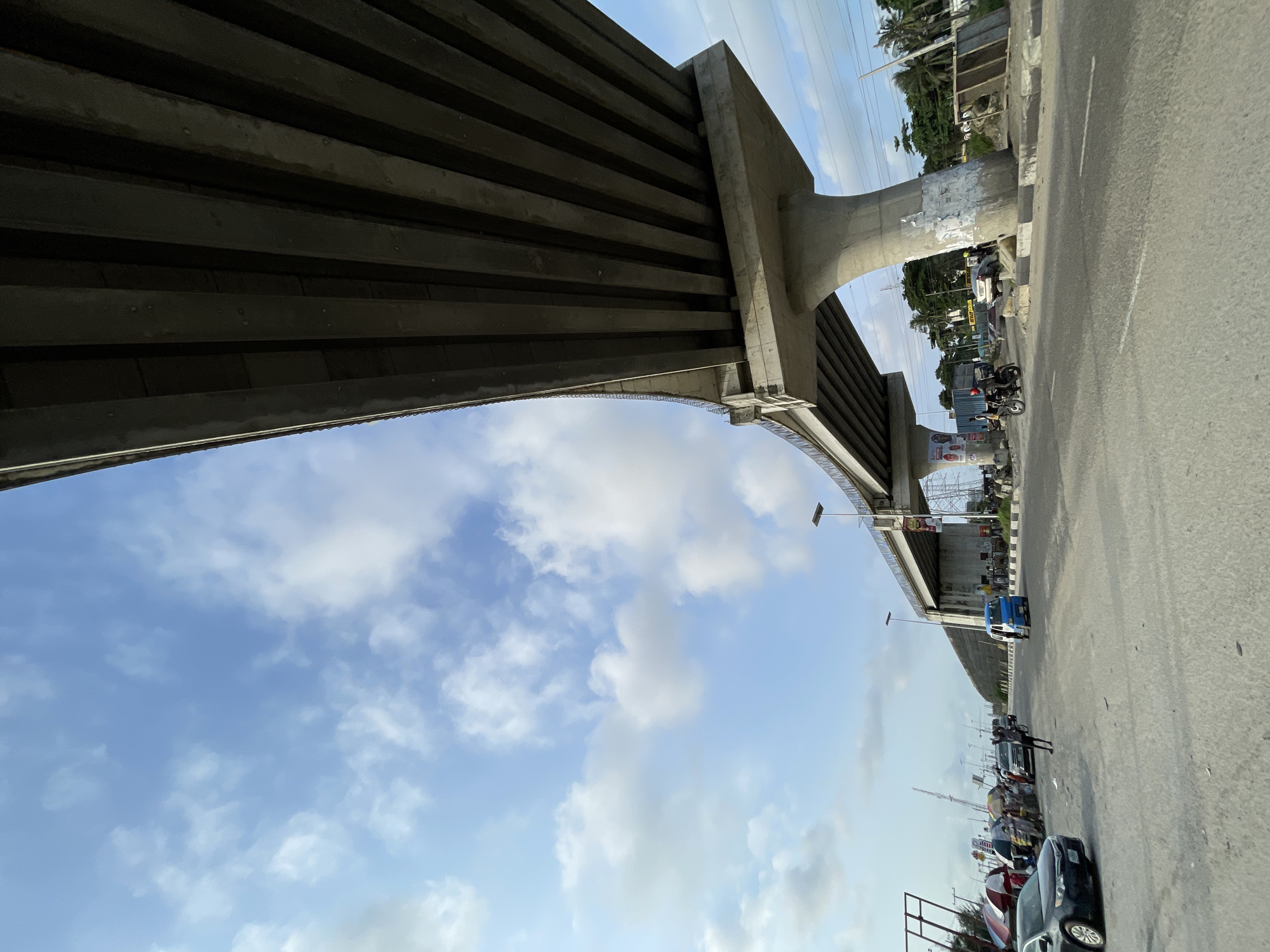 The image captures a view of an elevated highway under construction or recently completed in Lekki, Nigeria. The perspective is from below and to the side of the highway, looking up at its concrete structure. The highway is supported by large, curved concrete pillars. The sky above is a light blue with scattered white clouds. On the ground level, a road is visible with several cars and a blue minibus. People are gathered in small groups, some appearing to be vendors with umbrellas. A motorcycle with a rider is also present on the road. The overall environment suggests an urban setting, likely a busy area undergoing development.