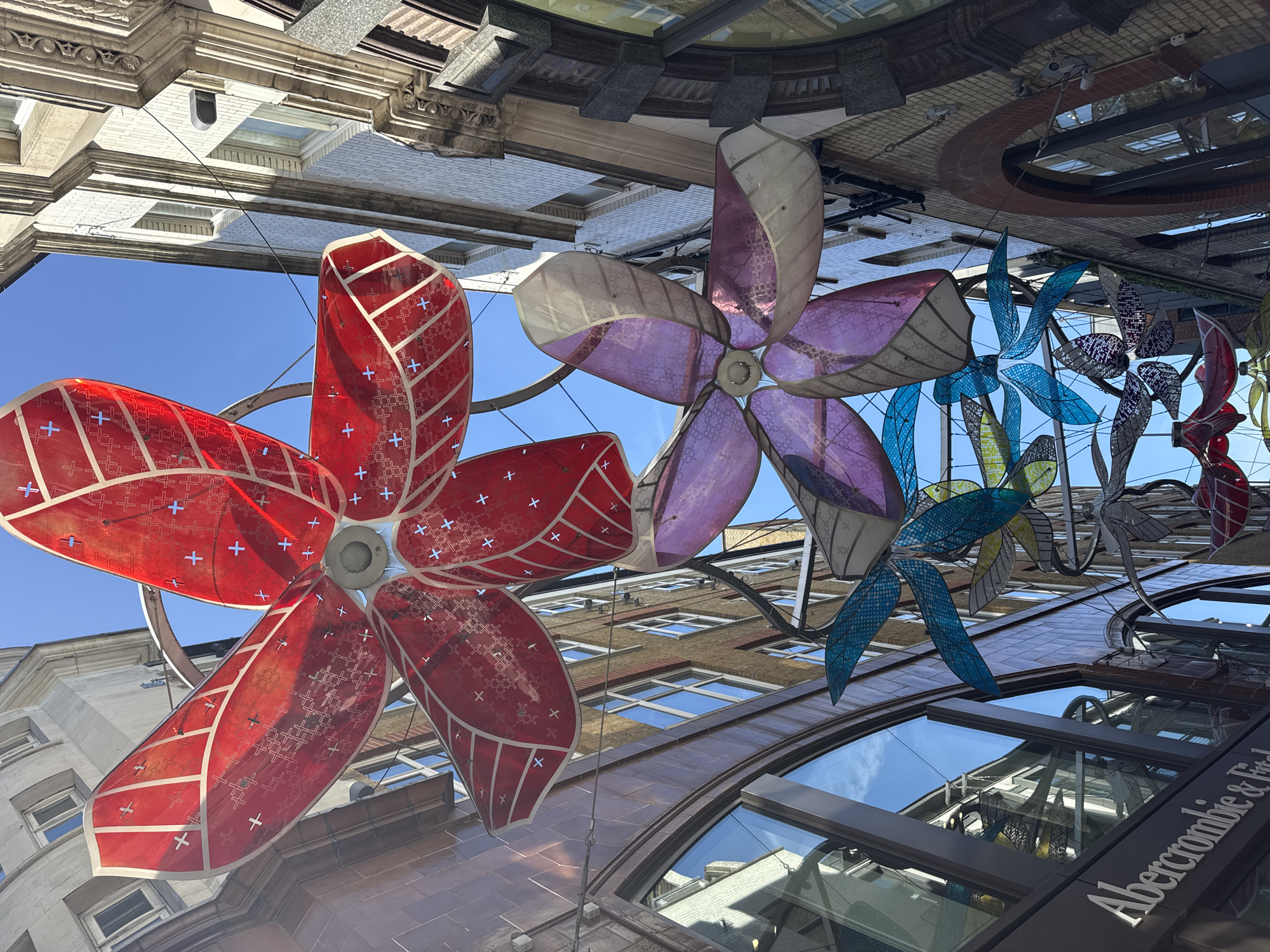 The image is a low-angle shot looking up at a street in London, United Kingdom, featuring colorful flower decorations suspended between buildings.
The focal point is the decorative flower installations in the center of the frame. There's a large red flower on the left, a light purple flower slightly behind it, and a series of blue, yellow, and red ornamental flower/leaf patterns toward the right. The buildings on either side consist of various architectural styles and facades, featuring windows, stonework, and architectural details.
An "Abercrombie & Fitch" store is present, as indicated by signage on a dark gray architectural element. The sky is visible between the buildings and decorations. The viewpoint is tilted.