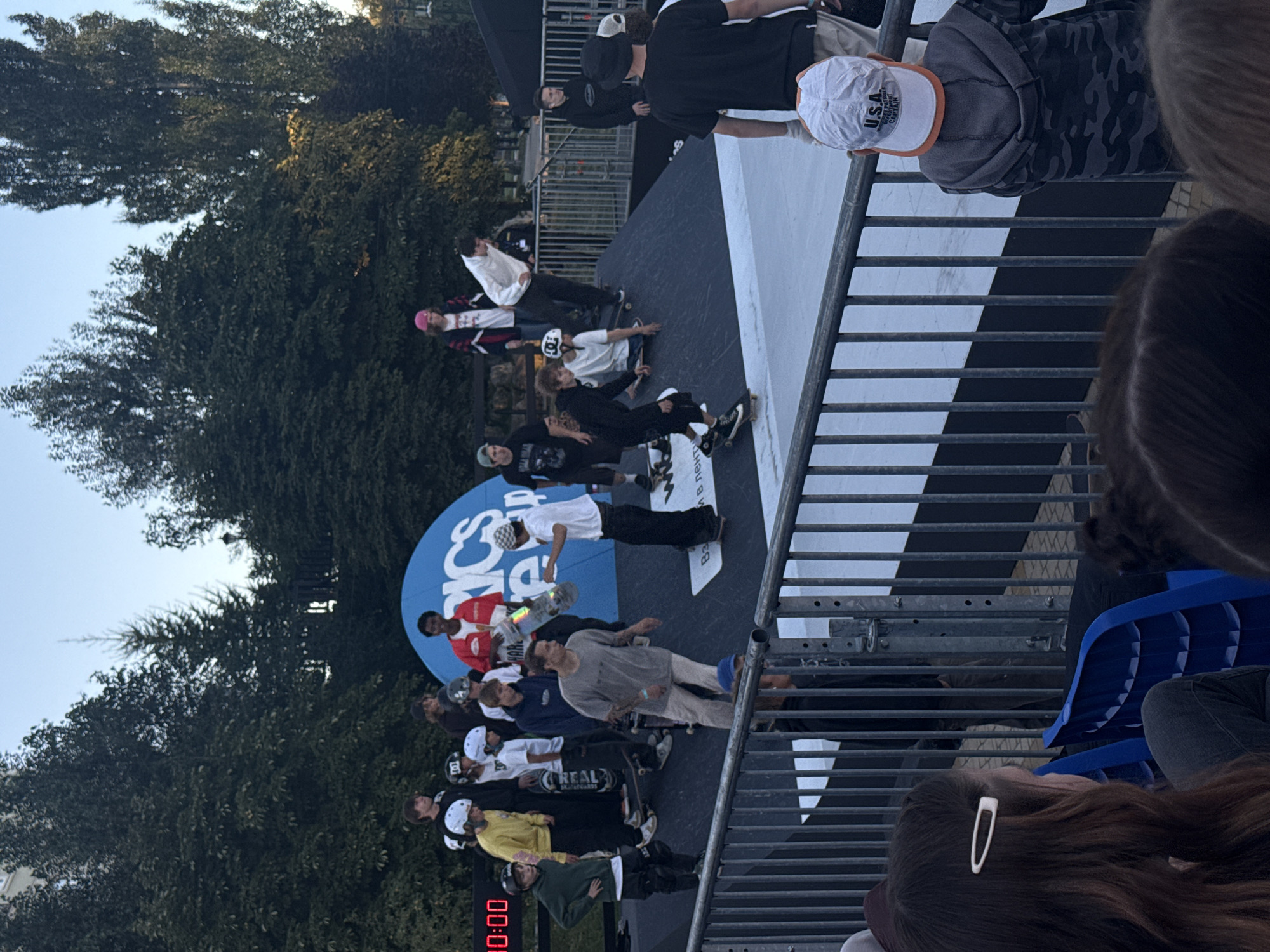 An outdoor skateboarding event is taking place in a park setting. A group of young people, mostly teenagers and young adults, are on a ramp structure. Several are skateboarding, some are standing and observing. One person in a red jacket is holding a skateboard. In the background, there are trees and a blue banner with "ASICS" logo. The time displayed on a digital clock in the background reads "00:00". The scene appears to be a public park with spectators in the foreground, seated in blue chairs behind a metal fence. The sky is clear and the lighting suggests it is late afternoon or early evening. The location is identified as the city of Ryazan, Russia, specifically in a park.