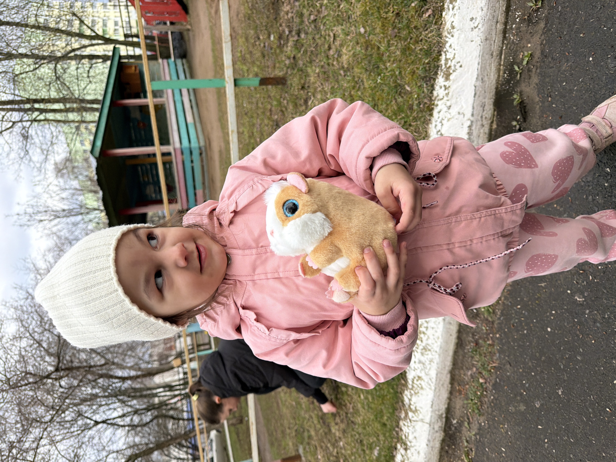 A young child is captured in an outdoor setting, likely a playground or park in Chaikovsky, Russia. The child, appearing to be a toddler or preschooler, is the central subject, looking slightly upwards and to their right with a soft, curious expression and a hint of a smile.
They are dressed in a light pink padded jacket, matching pink pants adorned with a repeating darker pink strawberry pattern, a white ribbed knit beanie, and light-colored sneakers or boots with star details. The child is holding a light brown and white stuffed animal resembling a guinea pig or hamster, which features prominent bright blue eyes.
The scene suggests an early spring day under an overcast sky, evidenced by the bare branches of trees forming the upper background. The ground is composed of a paved path with a white curb, bordering a patch of patchy green and brown grass. In the background, there is a colorful wooden playhouse-like structure with vertical slats in various hues of green, blue, pink, and red. Another person, partially visible in the mid-ground behind the child, is dressed in dark clothing and appears to be bending over near the grassy area. A light-colored building is faintly discernible through the bare tree branches in the distance. There is no visible text in the image.