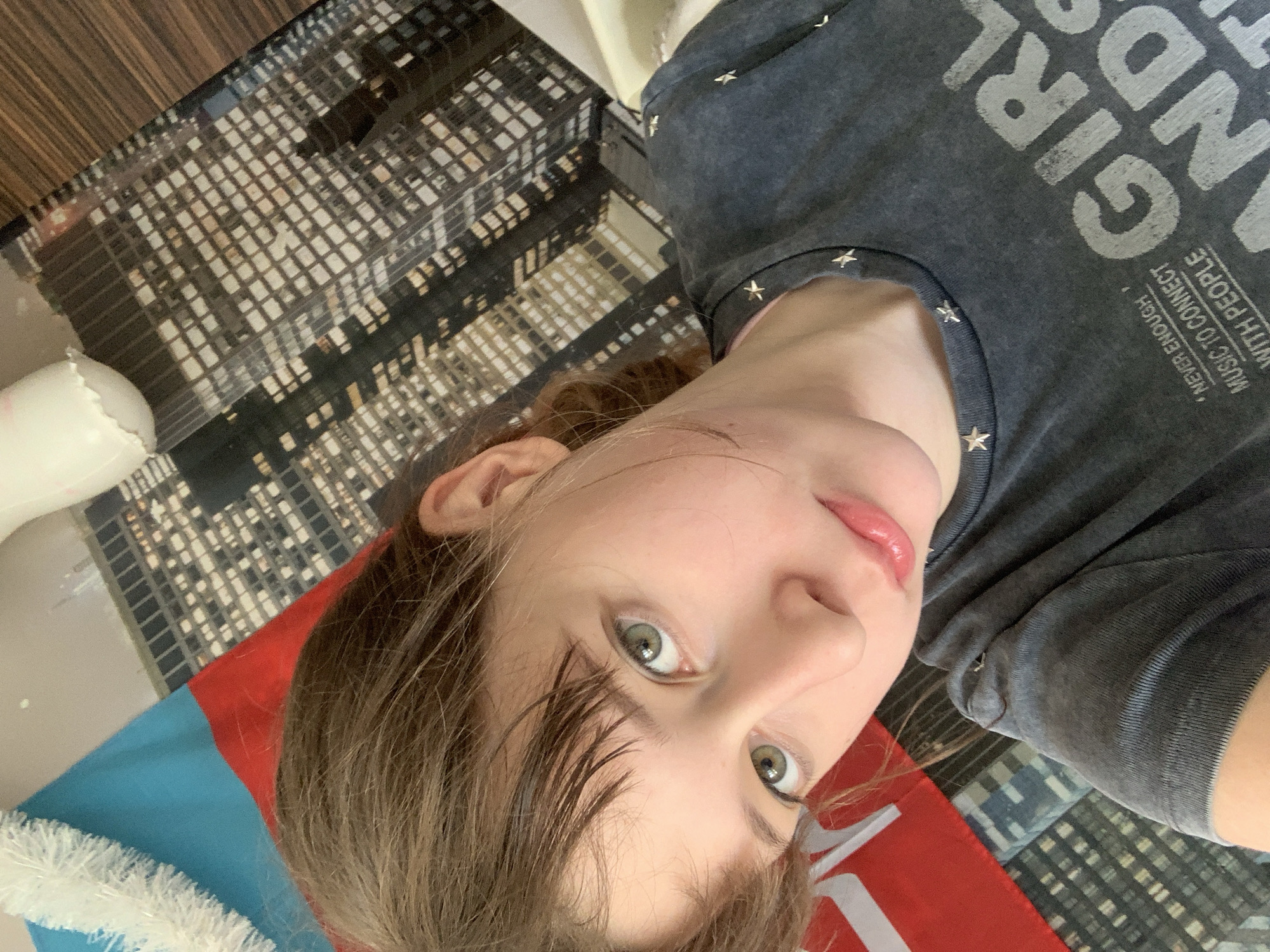A young person with light brown hair and green eyes is wearing a grey t-shirt with silver star studs around the neckline. The shirt has a printed design of the word "GIRLS". The background shows a floor with a pattern resembling city buildings, and a red, white, and blue object, possibly a flag. The location is not recognizable as Vozdovac Urban Municipality, Serbia based on the provided image.