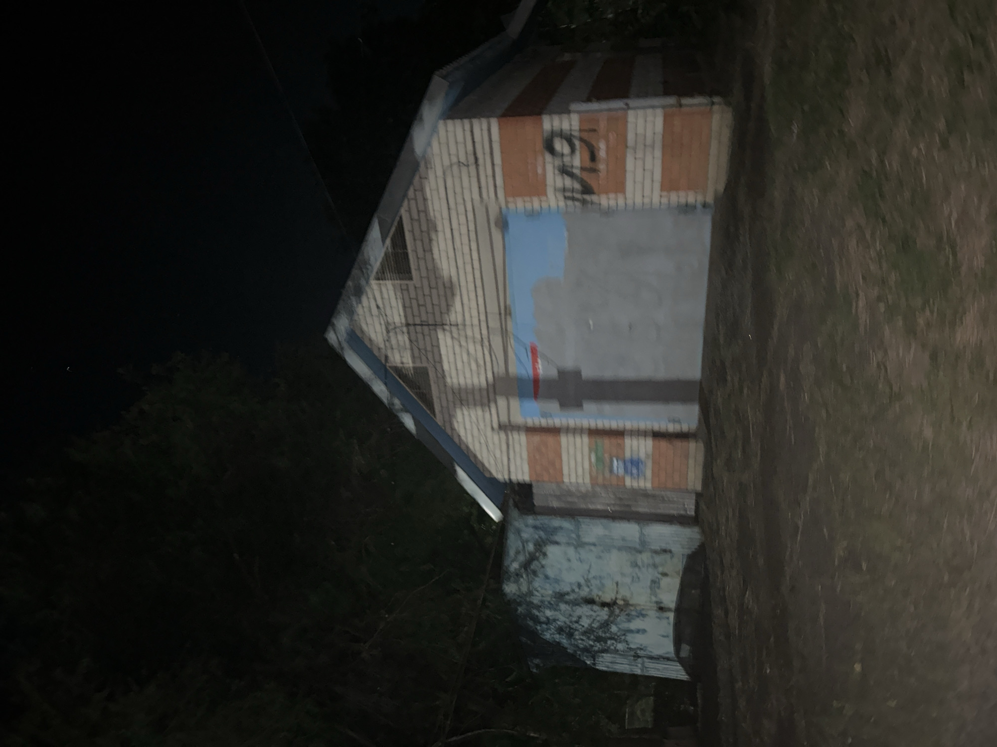 The image depicts a dimly lit outdoor scene at night, focusing on a garage with a blue and white striped metal door. The garage is constructed with light-colored bricks, interspersed with orange horizontal stripes. A blue and white striped structure is attached to the left side of the garage. In the background, dense foliage is visible, suggesting a natural or overgrown environment. The ground in the foreground appears to be dirt and grass. The overall impression is one of a somewhat neglected or rural setting. There are no people or discernible actions taking place in the image. The location is specified as Nevinnomyssk, Russia.