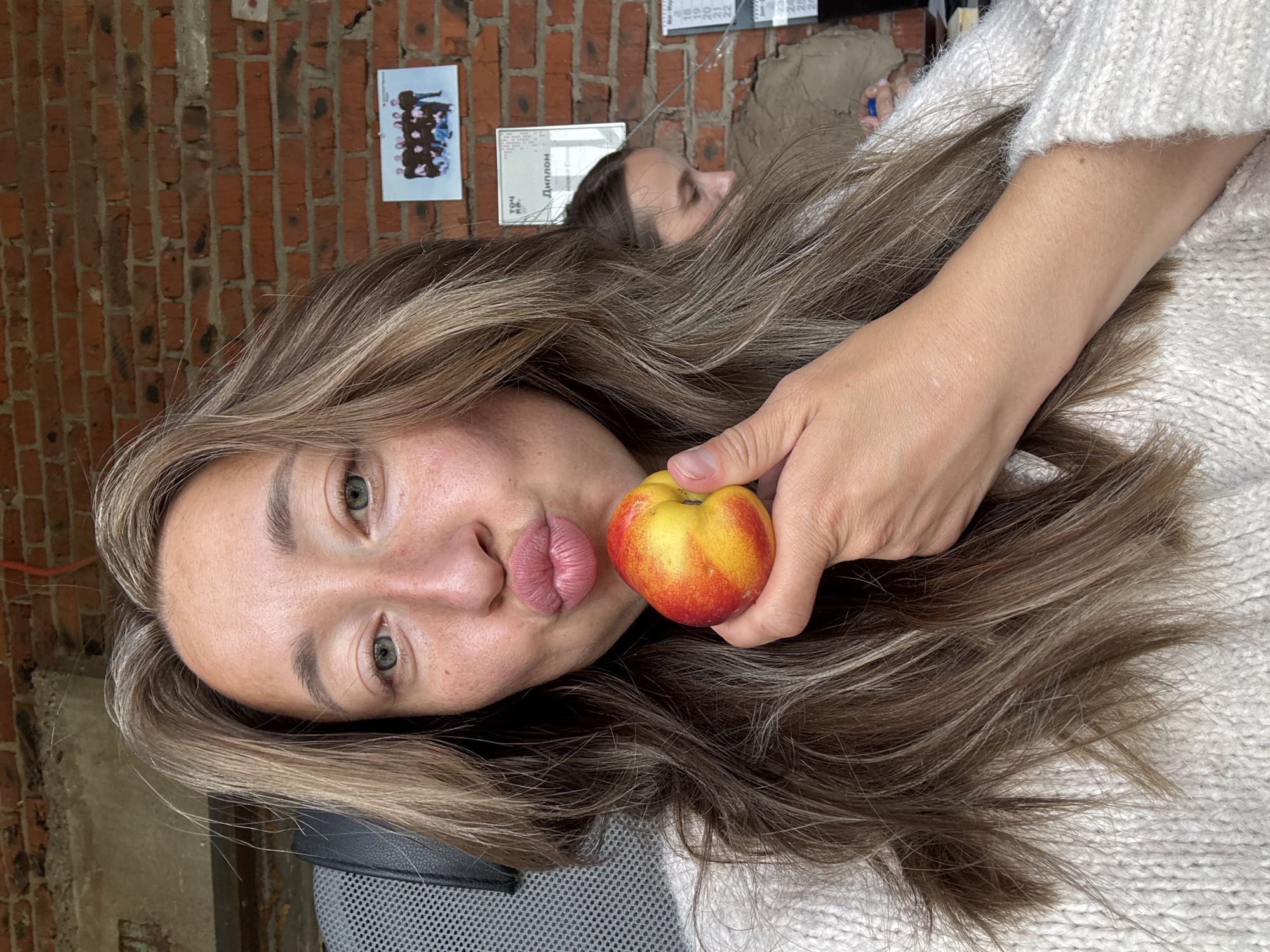 The image, appearing rotated 90 degrees clockwise, captures a young woman in a close-up selfie in an indoor setting, identified as Chaikovsky, Russia. She has long, light brown hair with blonde highlights and light-colored eyes. Her lips are puckered, and she holds a small, red and yellow fruit, resembling a nectarine or apple, in her right hand near her mouth. She is wearing a light-colored, possibly cream or off-white, knitted sweater.
In the blurred background, another person is partially visible, appearing to be resting or lying down. The primary background feature is a rustic red brick wall, to which several items are affixed. These include a small framed picture, likely a group photograph, and multiple documents. One document clearly displays the Cyrillic word "Диплом" (Diploma), indicating an academic or professional certificate. Another document, possibly a calendar or schedule, shows a sequence of numbers from "18" to "25". The lighting suggests it is daytime. The scene conveys a casual and playful mood, likely captured in a home, study, or office environment.