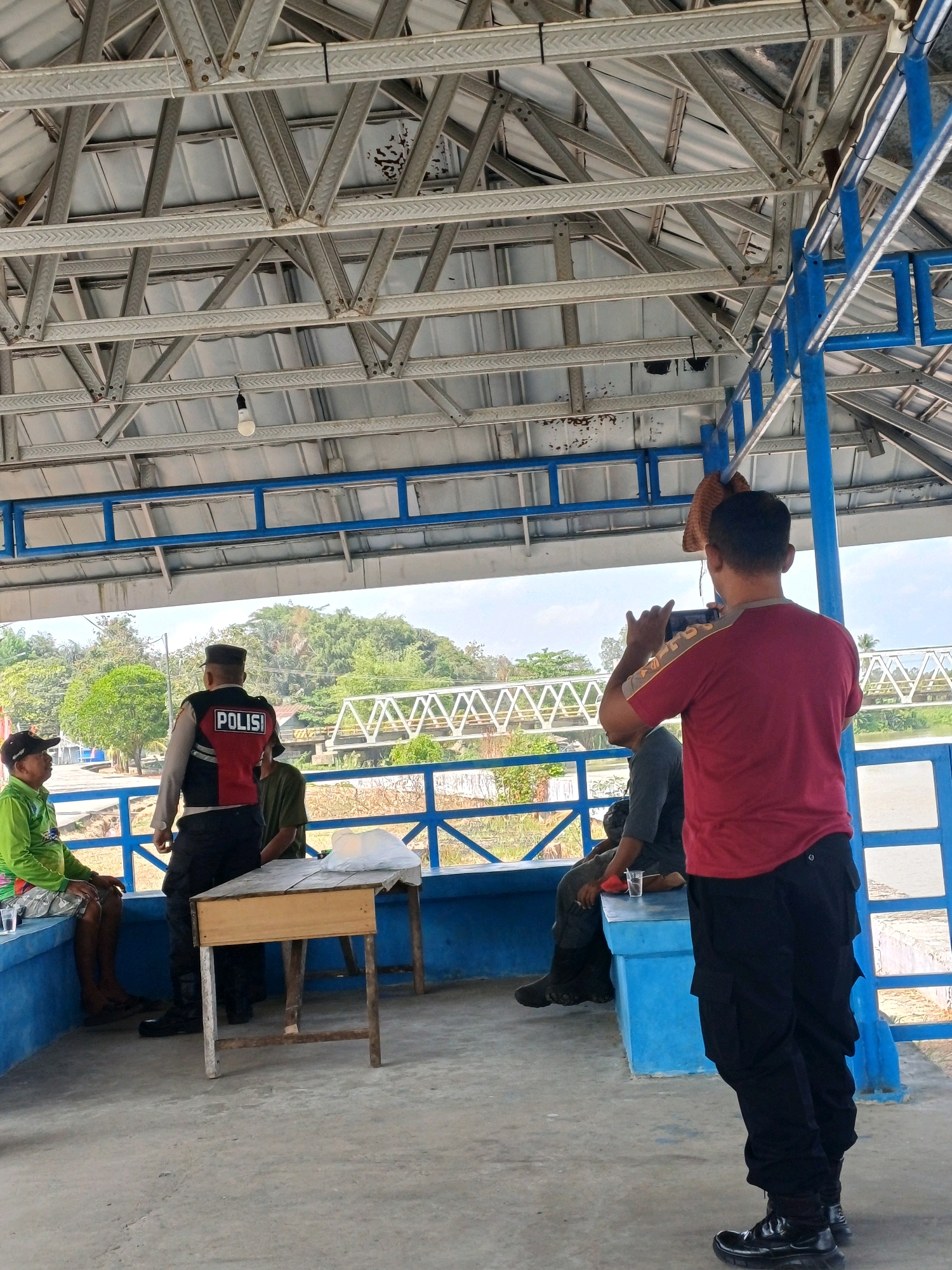 In Pasir Belengkong, Indonesia, a man in a red shirt and black pants stands facing away from the camera, holding a mobile phone to take a photo. He is positioned under a covered structure with a corrugated metal roof supported by a metal truss system. To his left, a police officer wearing a vest with "POLISI" on the back stands facing away from the camera. In front of the police officer is a small wooden table. To the left of the table, two individuals are seated on a blue bench; one is wearing a green long-sleeved shirt and patterned shorts, and the other is wearing a dark shirt. Behind them, a railing with blue bars runs along the edge of the covered area, overlooking a body of water and a treeline with a bridge in the distance. The ground is a concrete surface.