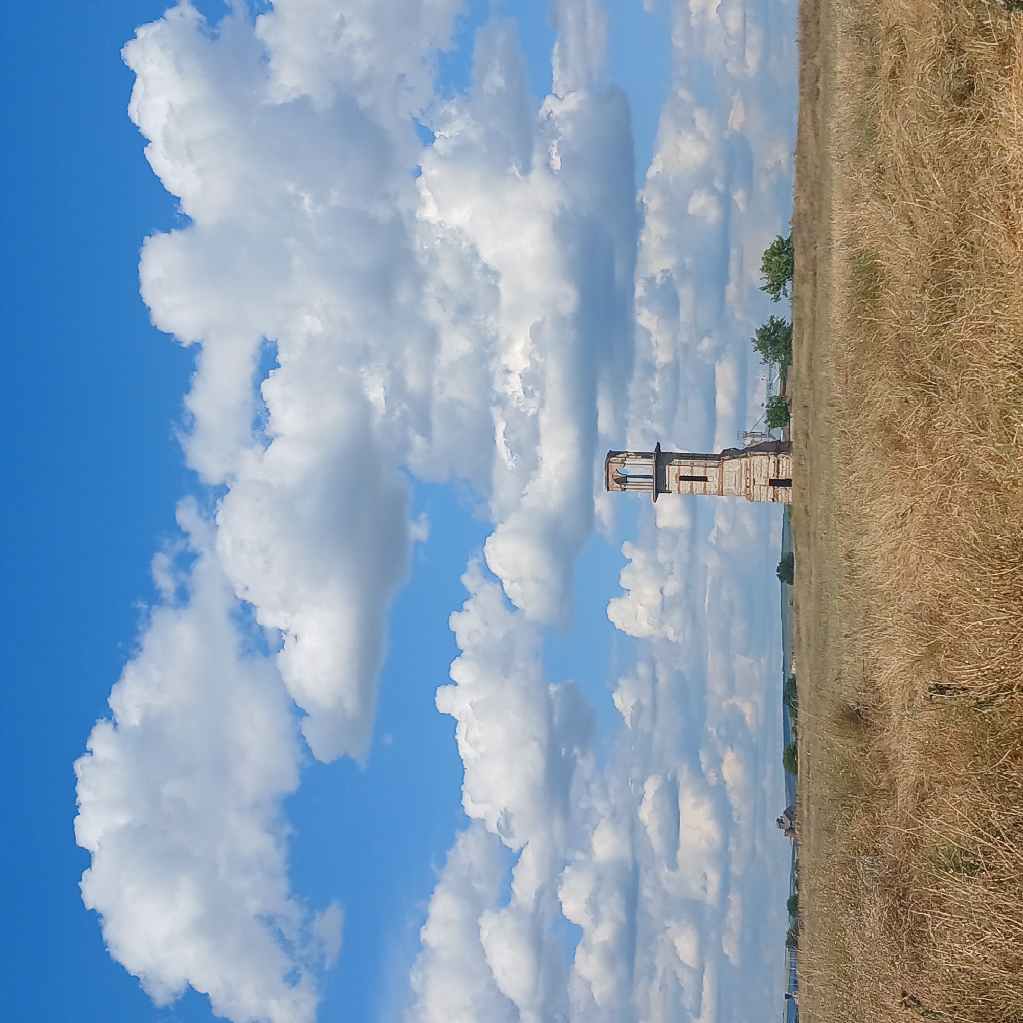 This is a photograph of a tall, dilapidated brick tower in a dry, grassy field under a bright blue sky with abundant white clouds. The tower, which appears to be an old church bell tower or watchtower, has arched openings at the top and shows signs of significant wear and tear, with exposed brickwork and crumbling sections. In the background, faint structures and trees can be seen on the horizon. The overall scene depicts a rural, somewhat abandoned landscape with a prominent, aging architectural element. The location is identified as Căzănești, Moldova.