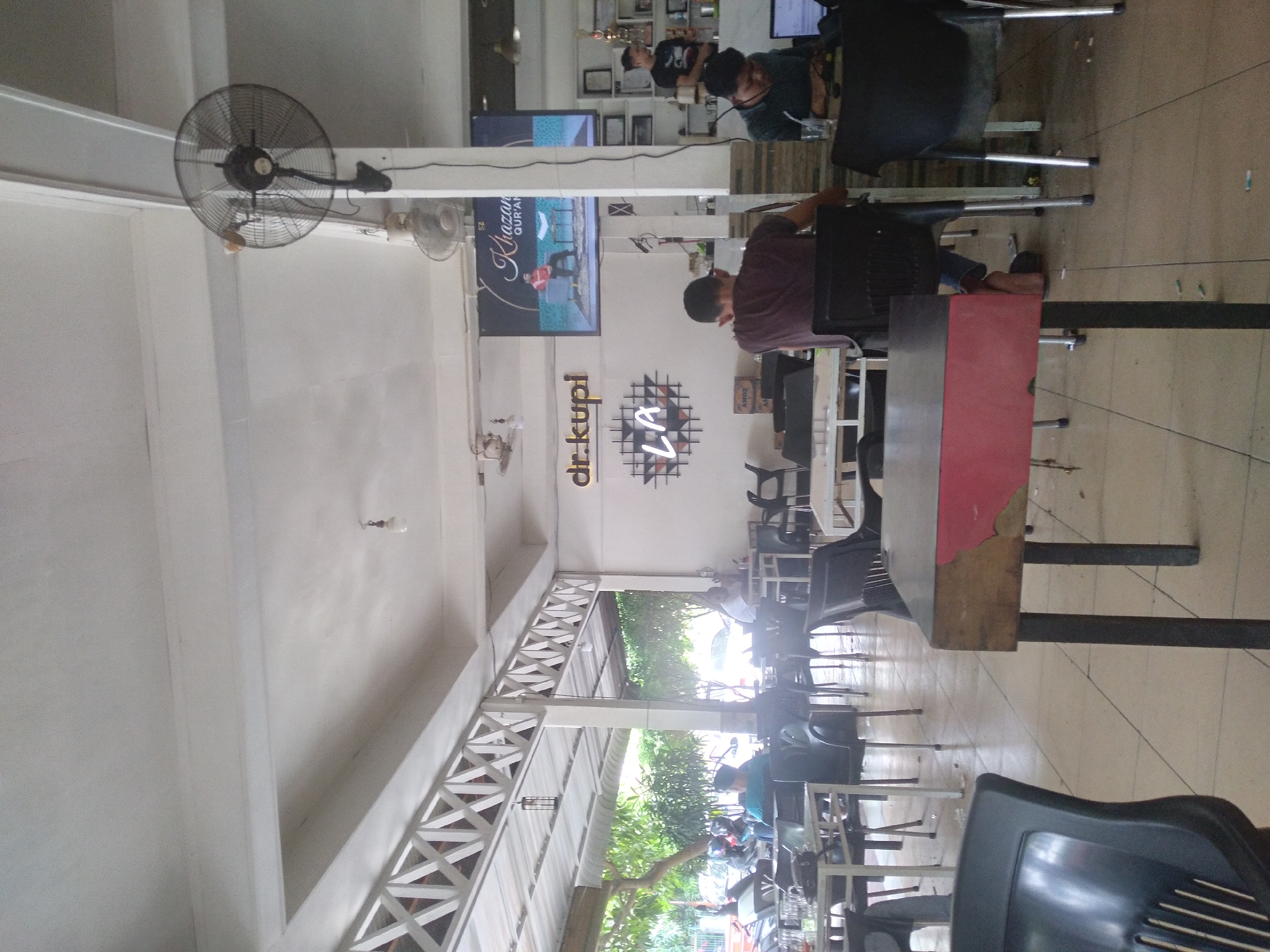 This media file captures the interior of a cafe or coffee shop, partially open to an outdoor area, likely during the daytime. The setting features white walls, a light-colored tiled floor, and a decorative white lattice beam structure separating the indoor seating from a greener, semi-outdoor section with trees.
Several individuals are present, mostly men. In the foreground on the right, a person in a dark, possibly maroon, shirt is seated at a table with a prominent red and gray tabletop. Further back, another person in a dark top is visible behind a table, appearing to focus on a screen. In the background, another individual in a black shirt is seated at a table.
The cafe is furnished with dark plastic chairs with metal legs and various tables. Several wall-mounted fans are visible, one prominently in the upper left, indicating a need for ventilation. A large television screen on the wall displays text that reads "Khazanah Qur'an," suggesting a program related to Islamic knowledge. Below the screen, the cafe's branding is visible: "dr.kupi" in a prominent font, alongside a stylized "LA" within a geometric pattern.
The outdoor area shows more seating and hints of vehicles, possibly motorcycles, indicating that it's a bustling public space. The overall atmosphere appears casual and relaxed, with people engaged in typical cafe activities such as working, socializing, or watching the television.