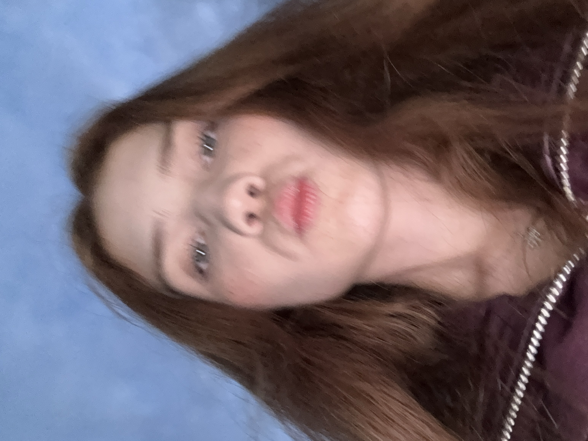 A close-up, low-angle portrait depicts a single individual, appearing to be a young woman, directly facing the camera. She has long, reddish-brown hair parted near the center, framing her face. Her lips are pursed in a "kissing" or "duck face" expression. Her eyes are open, looking directly forward, with a light reflection visible in each pupil. She has a fair complexion with a subtle reddish tint on her cheeks. The subject is wearing a dark, possibly maroon or purple, outerwear garment, featuring a metallic zipper whose teeth are visible across the lower edge of the frame. The background is a solid, uniform light blue color, devoid of any discernible features or objects. The image exhibits a slightly soft focus, consistent with a self-taken photograph. No identifying information regarding location or specific environment is present.