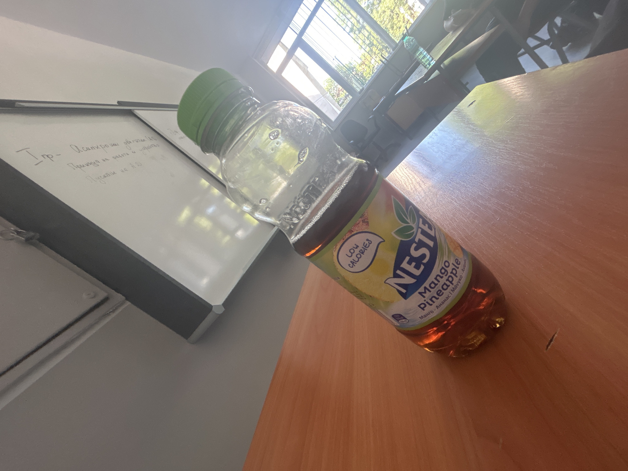 A clear plastic bottle containing a brownish liquid with some bubbles is placed on a light brown wooden table. The bottle has a green cap and a label that reads "Nestea Mango Pineapple" and "LOW CALORIES". In the background, a whiteboard with some writing in a foreign language is visible, along with a window showing trees and sunlight. The scene appears to be indoors, possibly in a classroom or office. The camera angle is from above, looking down at an angle.