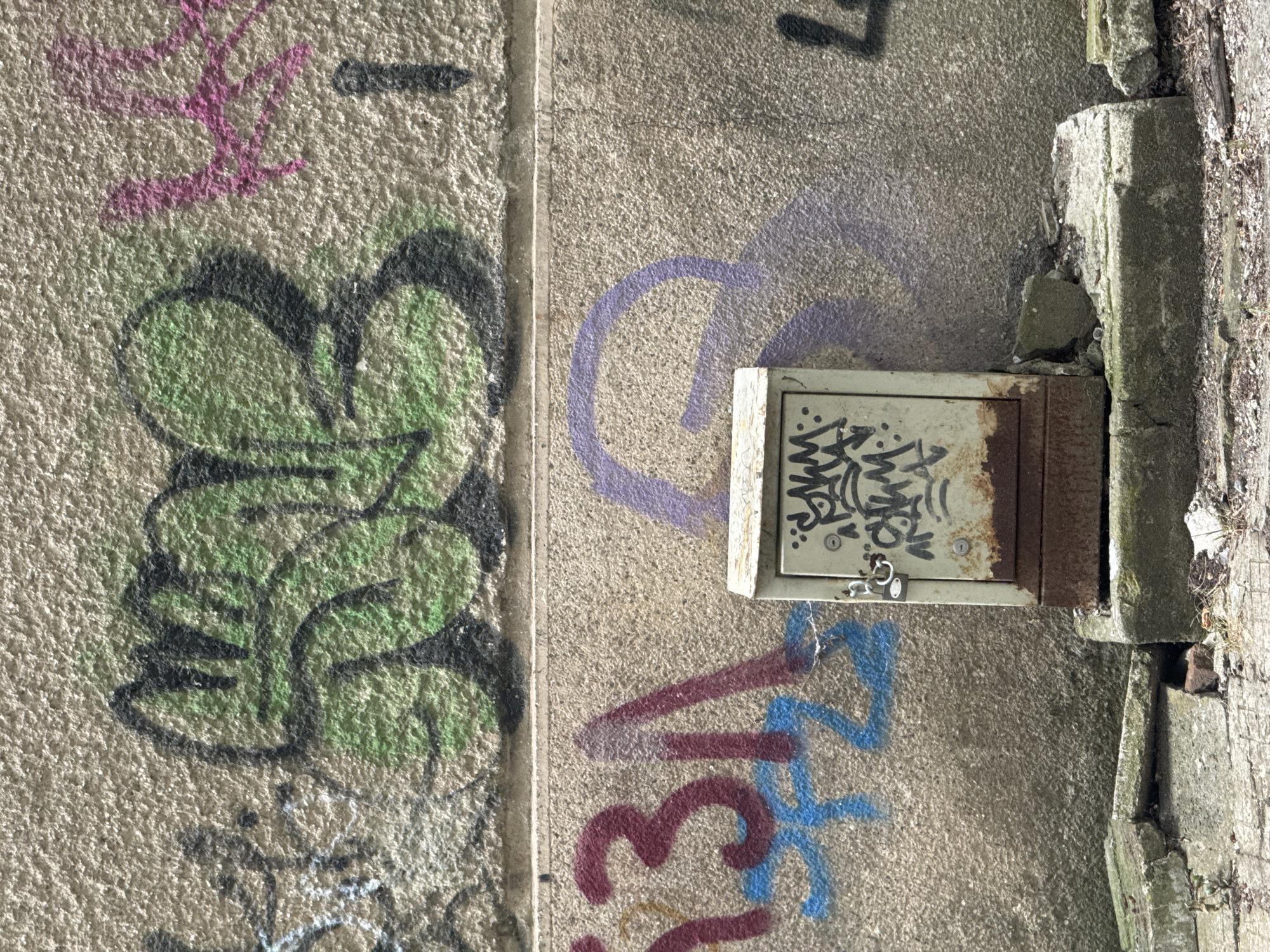 The image shows a close-up of a concrete wall with graffiti and a small metal box. The wall is textured and appears aged. In the upper section of the wall, there is graffiti in green and black with some pink markings to the right. The lower section of the wall features larger graffiti in burgundy and blue, with a purple outline of a shape above the metal box. The metal box is grey and has rust stains at the bottom. It is locked with a silver padlock and has some black graffiti tags on its front. The box is positioned on a concrete base that is partially broken. The ground in front of the base is paved with concrete slabs, some of which are cracked and broken, with debris scattered around. The scene suggests an urban environment, possibly an alleyway or the side of a building. This image was taken in the city of Sofia, Bulgaria. There are no people present in the image.