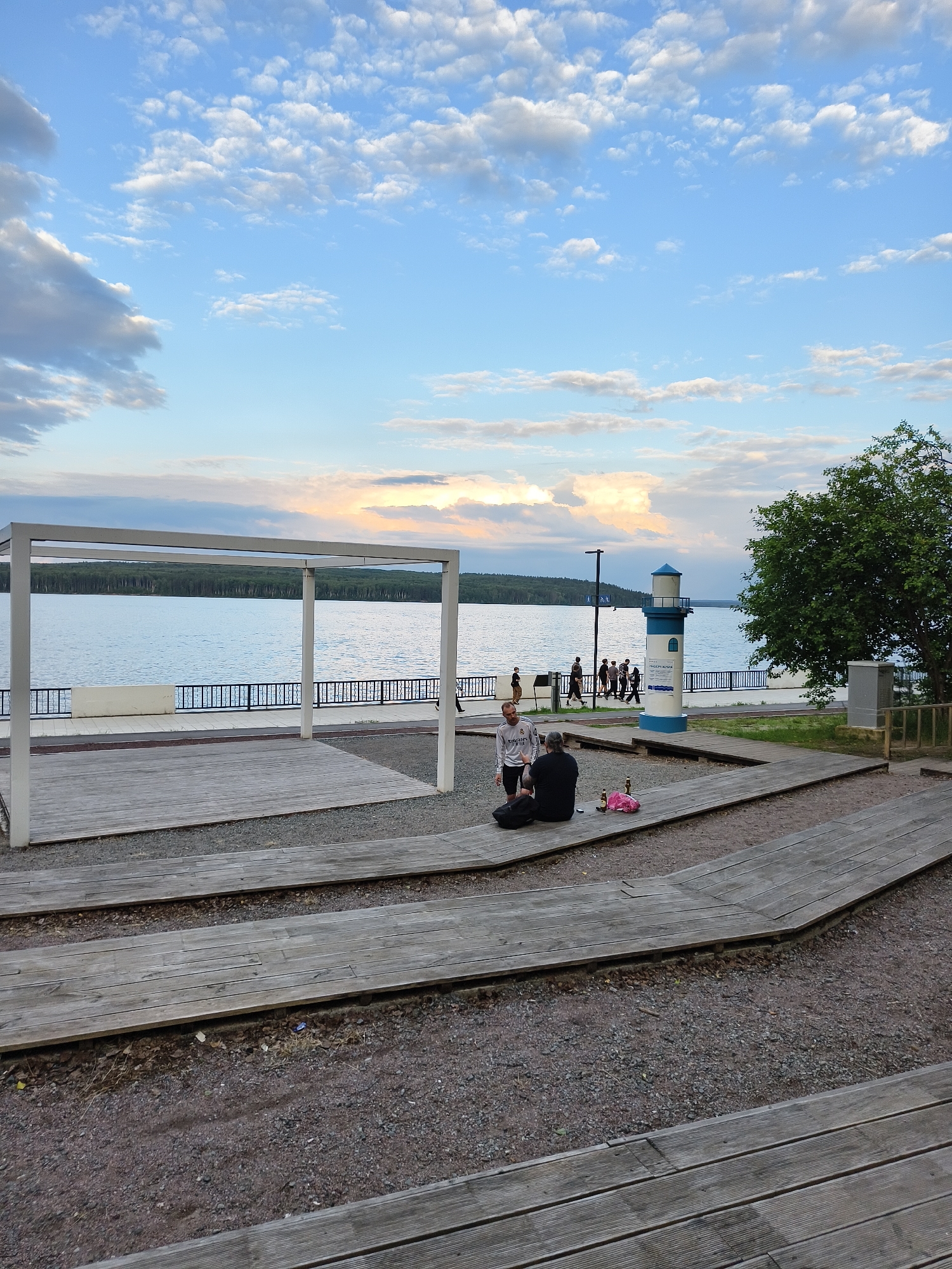 This media file depicts a scenic view of a waterfront area with a cloudy sky. The scene features a large body of water, likely a river or lake, bordered by a treeline on the opposite shore. In the foreground, there are wooden tiered seating areas constructed with wooden planks, set against a gravel ground.
Several people are present in the image. Two individuals are sitting on the wooden seating in the foreground, engaged in conversation or relaxing. A small group of people are walking along a paved path adjacent to the water. Further down the path, a small lighthouse-like structure with blue and white stripes stands prominently. Some people are gathered near this structure.
The time of day appears to be late afternoon or early evening, indicated by the soft lighting and the colors in the sky, which show hues of blue, white, and hints of orange and pink from the setting sun. The weather seems pleasant, with scattered clouds creating a dynamic sky.
Notable details include the architectural elements such as the white rectangular shelter in the mid-ground, the railing along the waterfront, and the lighthouse. A sign is visible on the lighthouse, though the text is not clearly legible in this resolution. The overall mood of the scene is relaxed and tranquil, suggesting a public park or recreational area. The location context of Chaikovsky, Russia, further grounds the scene to a specific geographical setting.