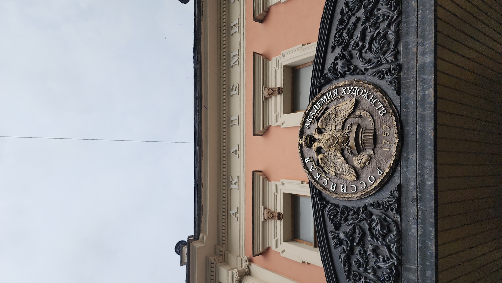 The image shows the Russian Academy of Arts in Saint Petersburg, Russia. The building is peach-colored with white trim. A bronze plaque with a double-headed eagle and the inscription "Российская Академия Художеств 1757" (Russian Academy of Arts 1757) is mounted on a black gate. The sky is overcast. The building's facade includes window details and carved ornaments.