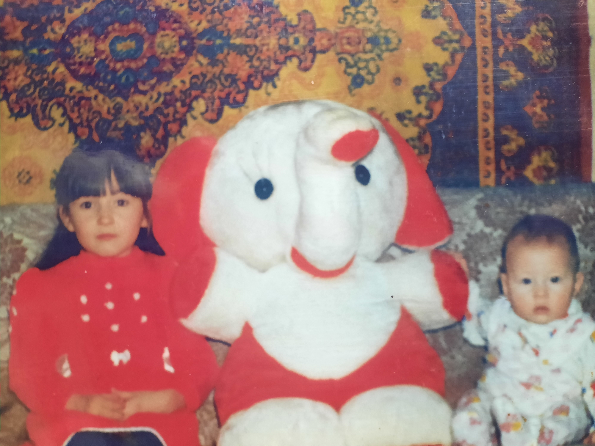 The image captures a still shot featuring two children and a large stuffed elephant, likely taken indoors.
On the left, a young girl with straight, dark hair and bangs wears a red sweater with white polka dots and a bow detail. Her hands are clasped in front of her. In the center, the oversized stuffed elephant is predominantly white with red accents on its ears, trunk, and chest. It has black button eyes. To the right, a baby sits wearing patterned pajamas.
The background is dominated by a tapestry or rug with complex, colorful patterns. The setting gives no definitive clues to its location in Архангельское, Russia. The environment is clearly someone's home.