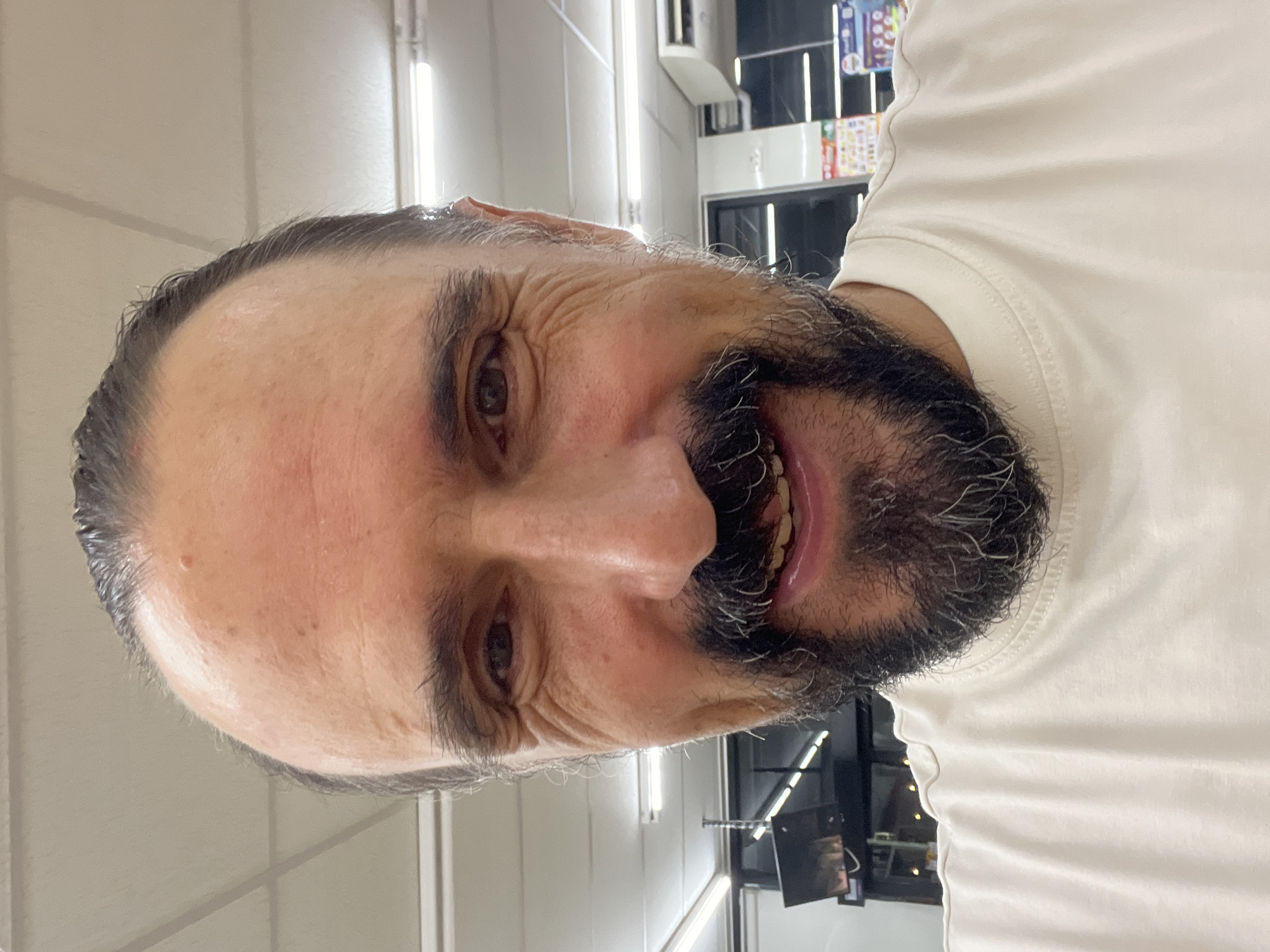 A close-up selfie of a middle-aged man with dark hair pulled back, a receding hairline, dark eyebrows, and a full beard with streaks of gray. He has brown eyes and is smiling, revealing his teeth. He is wearing a light-colored crew neck t-shirt. The background appears to be indoors, with fluorescent lighting visible on the ceiling. The man's face and upper chest fill the frame. The image does not provide any specific details about the location in the city of บ้านบางรักษ์, Thailand.