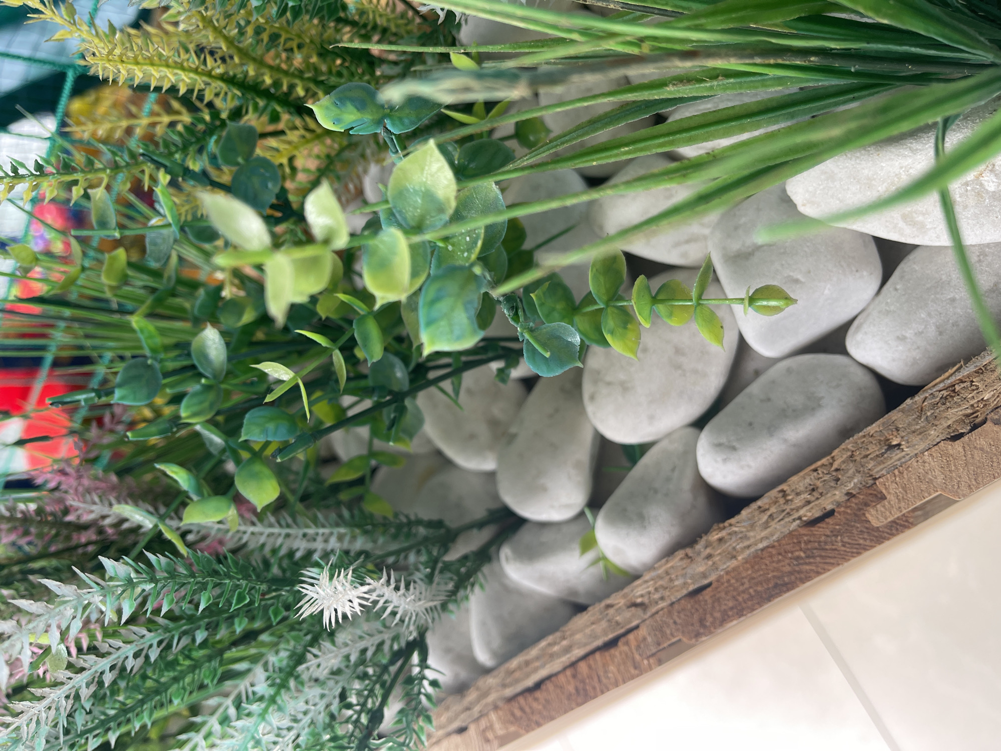 An artificial plant arrangement is displayed in a weathered wooden planter. The planter contains numerous smooth, white stones, creating a ground cover. Several types of artificial plants are arranged within the stones, including greenery with small, rounded leaves, feathery ferns in shades of green and white, and spiky yellow-green foliage. The composition is dense and varied, suggesting a decorative display. The background is blurred, but hints of red and green suggest other decorative elements or a colorful environment. The image is taken from a close-up perspective, focusing on the texture and detail of the plants and stones. No people or discernible location in a city are present.