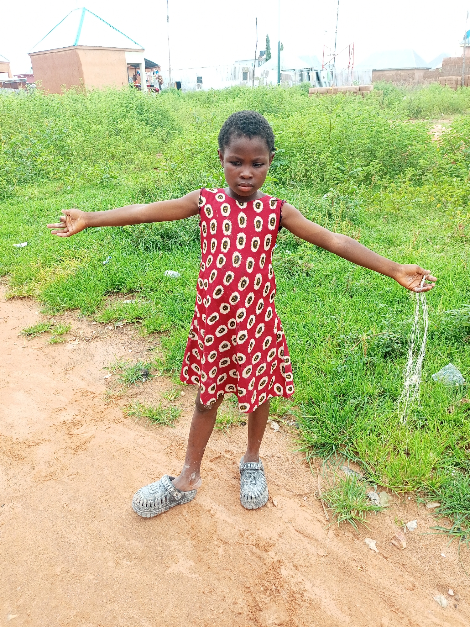 A young Black girl stands in a dirt and grass area, facing forward with her arms extended outward. She is wearing a red dress with a white circular pattern and grey clogs. She is holding a bundle of white strings in her right hand. In the background, there are buildings and green vegetation. The location is identified as the city of Gombe, Nigeria.