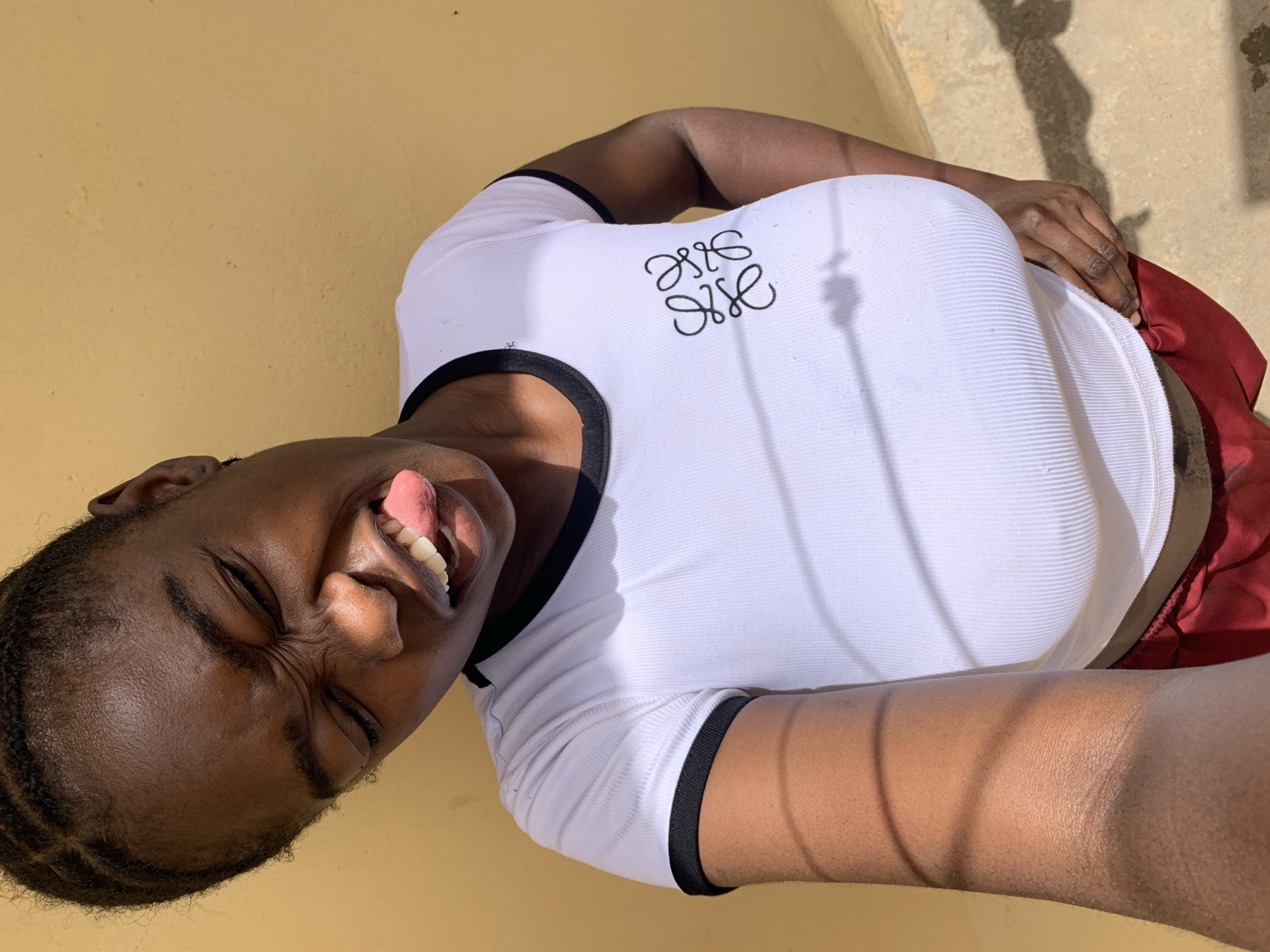 A medium shot shows a woman with dark skin, brown eyes, and short braids. She is outdoors and wearing a white, short-sleeved top with a black trim around the collar and sleeves. The shirt has a stitched black logo on the upper left. She also wears maroon bottoms. She is sticking out her tongue and squinting, and appears to be smiling. The background consists of a yellow, slightly textured wall, and a concrete walkway with a faint shadow. The location may be in or near Rafin Par, Nigeria, based on the user's request.