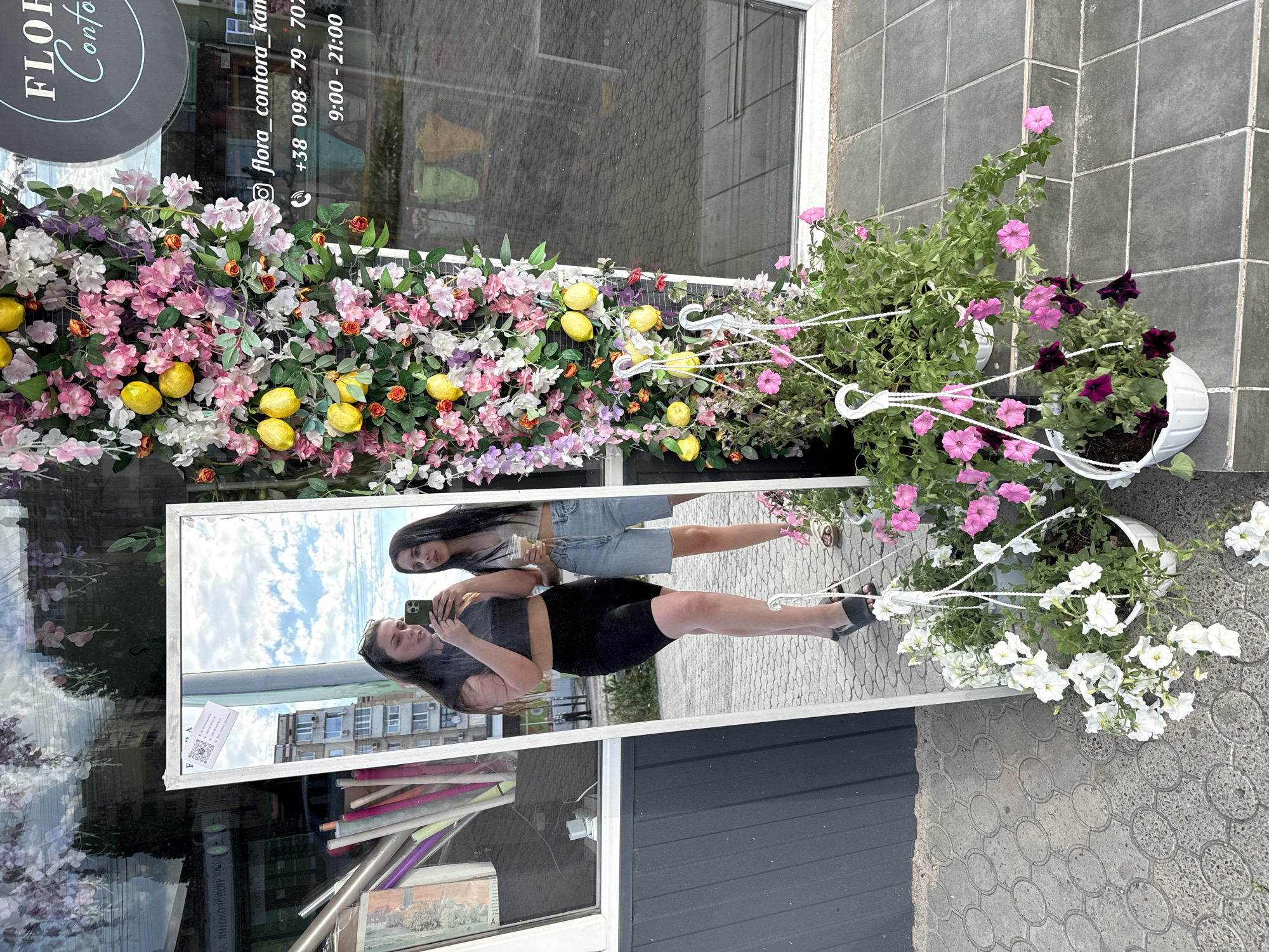 This is a vertically oriented photograph of a storefront decorated with artificial flowers and lemons. In the foreground, a mirror reflects the scene, showing two young women. One woman in the reflection, wearing a black top and black shorts, is holding a phone to take a selfie. The other woman, partially visible, wears a grey top and denim shorts and holds a drink. The storefront window displays floral arrangements, and text on the window reads "FLOR Conto" and includes a phone number "+38 098-79-702" and business hours "9:00 - 21:00". White planters filled with pink and white petunias hang to the right of the storefront. The ground is paved with patterned tiles. The scene appears to be outdoors during the daytime, with a cloudy sky reflected in the mirror.