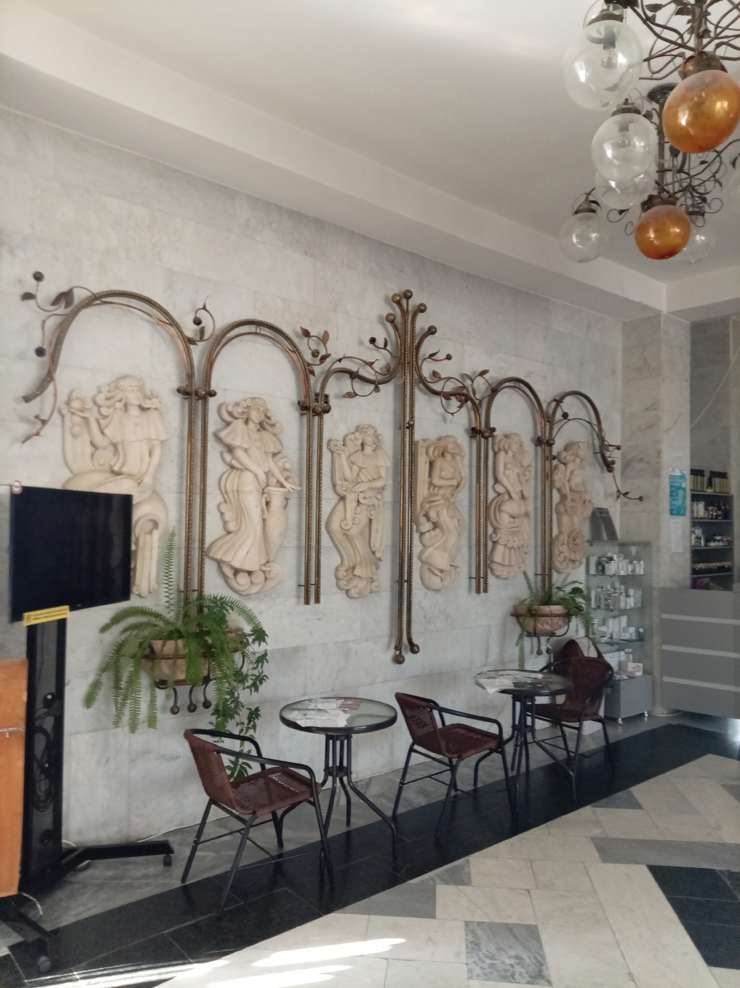 The image depicts an interior scene, likely a waiting area or lobby, featuring a marble-tiled wall adorned with decorative metal arches and bas-relief sculptures of female figures. In the foreground, there are two small tables with chairs, one with a glass top and the other empty. A large potted fern is visible, and to the left, a black screen is mounted on a stand. The floor is a mosaic of marble tiles in black, white, and grey. In the background, a glass display cabinet filled with bottles and toiletries suggests a commercial or spa setting. The lighting appears to be artificial, with a chandelier featuring glass and amber-colored globes hanging from the ceiling. The overall impression is one of an elegantly decorated indoor space. No people are visible in the image. The location is identified as the sanatorium "Slavyanovskiy" in Zheleznovodsk, Russia.