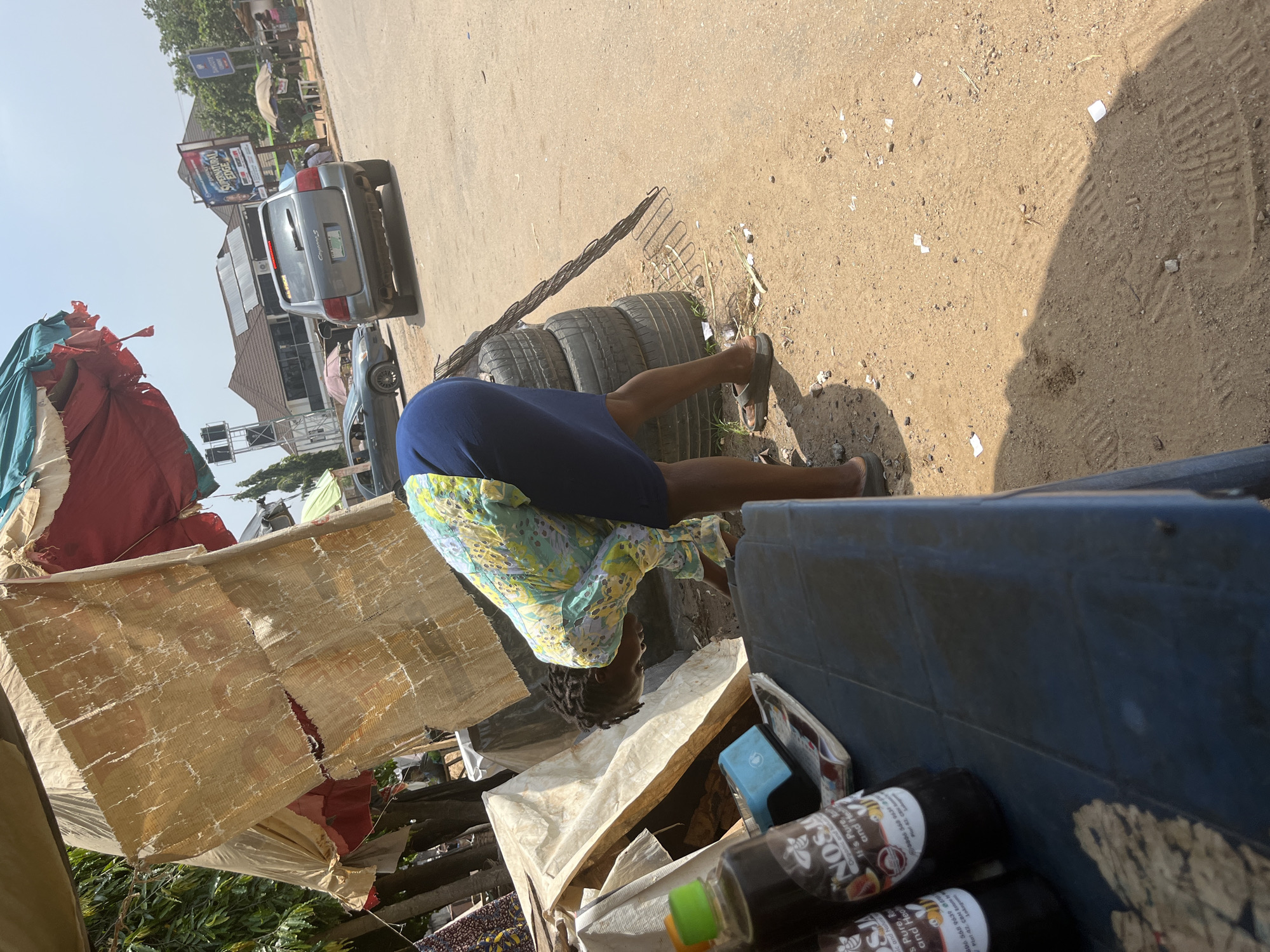 The image captures an outdoor scene in Abuja, Nigeria, likely near a roadside market. In the foreground, a person wearing a colorful floral-patterned shirt and a dark blue skirt is bending over, appearing to sort through items. Their hair is braided, and they are wearing sandals. To the left, a blue plastic table holds two bottles of what appears to be honey, labeled "NOSJ Organic and Natural." Behind the person, a stack of old tires is visible. The environment is dusty and appears to be a street with a paved road in the background. Several vehicles, including a silver SUV and a darker sedan, are on the road. Overhead, a makeshift stall with a torn fabric canopy provides shade. The scene is illuminated by bright sunlight, casting shadows. A few scattered pieces of white paper are visible on the ground.