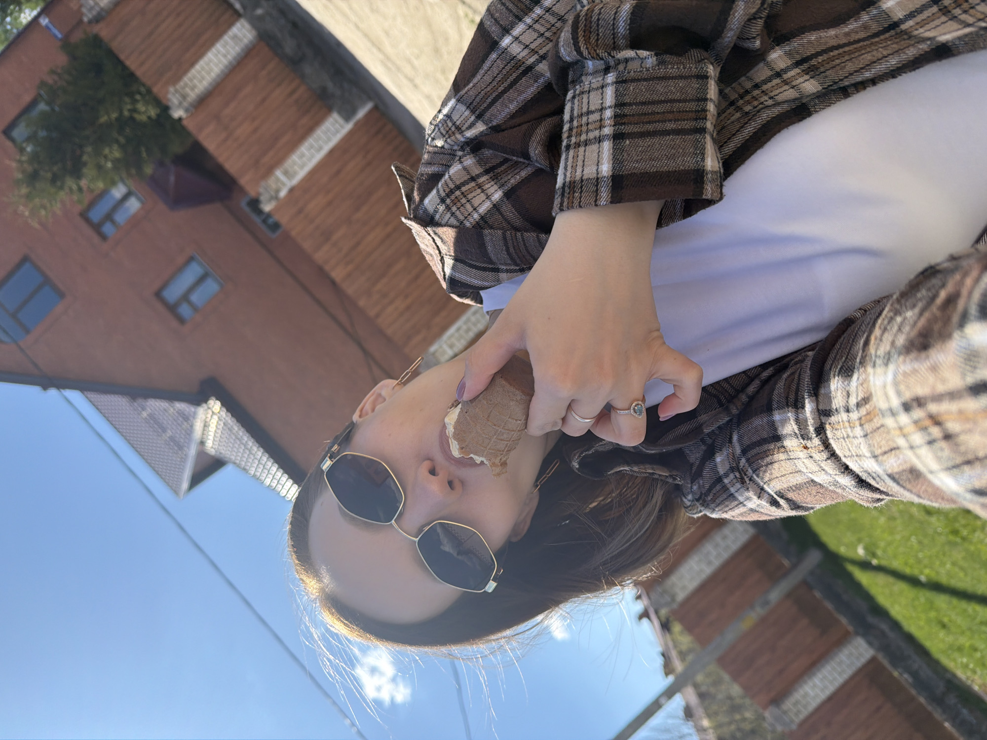 A woman is holding an ice cream cone to her mouth, against an outdoor setting in Reshetnikovo, Russia. She's wearing sunglasses, a white top, and a brown plaid jacket. She is also wearing rings on her fingers. In the background, a building with a brown roof, trees, and a blue sky are visible. The scene is captured at an angle, suggesting the picture was taken as a selfie.