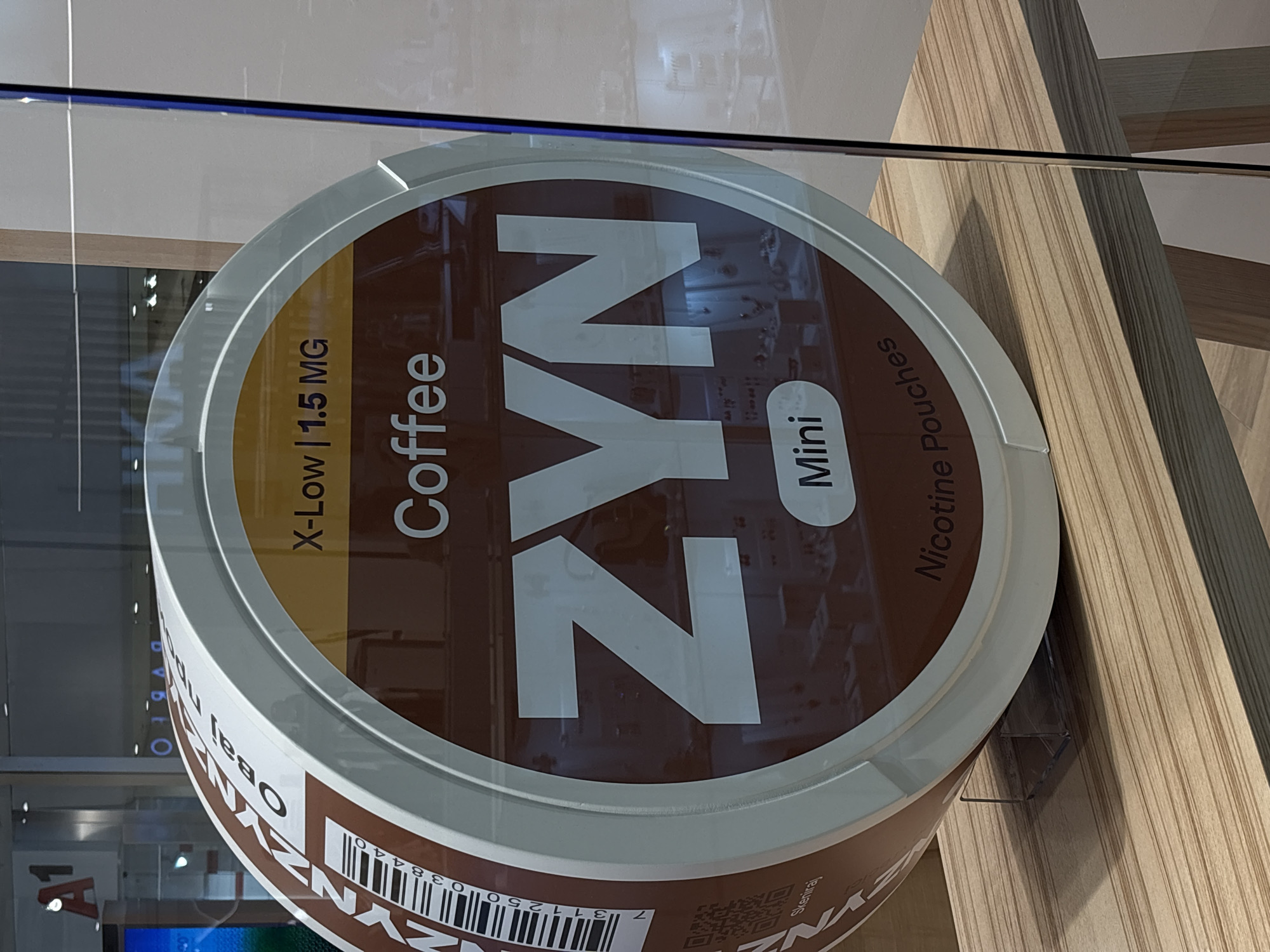 The image displays a large, cylindrical promotional item, designed to look like an oversized container of ZYN nicotine pouches, positioned behind a clear glass panel. The primary colors of the display are brown and white.
The top surface of the cylindrical object prominently features the brand name "ZYN" in large white letters. Other discernible text on the lid indicates the product details: "Coffee" for the flavor, "X-Low | 1.5 MG" for the nicotine strength, and "Mini" for the pouch size, followed by the description "Nicotine Pouches."
The background shows reflections on the glass, suggesting an indoor setting with artificial lighting. To the right, a modern architectural element made of light-colored wooden paneling is visible.
On the side of the cylindrical display, a partial barcode with the number "311250038440" is present. Below the barcode, the word "Skeniraj," which is Serbian for "Scan," is visible, along with a QR code. Additionally, fragments of Serbian text, "Овај пр," appear, likely part of a phrase like "Овај производ" (This product).
No people are present, and no specific activity is taking place; the image captures a static product display. The indoor setting, potentially a store or exhibition, is brightly lit, but the time of day is not determinable. The image was taken in Kragujevac, Serbia.