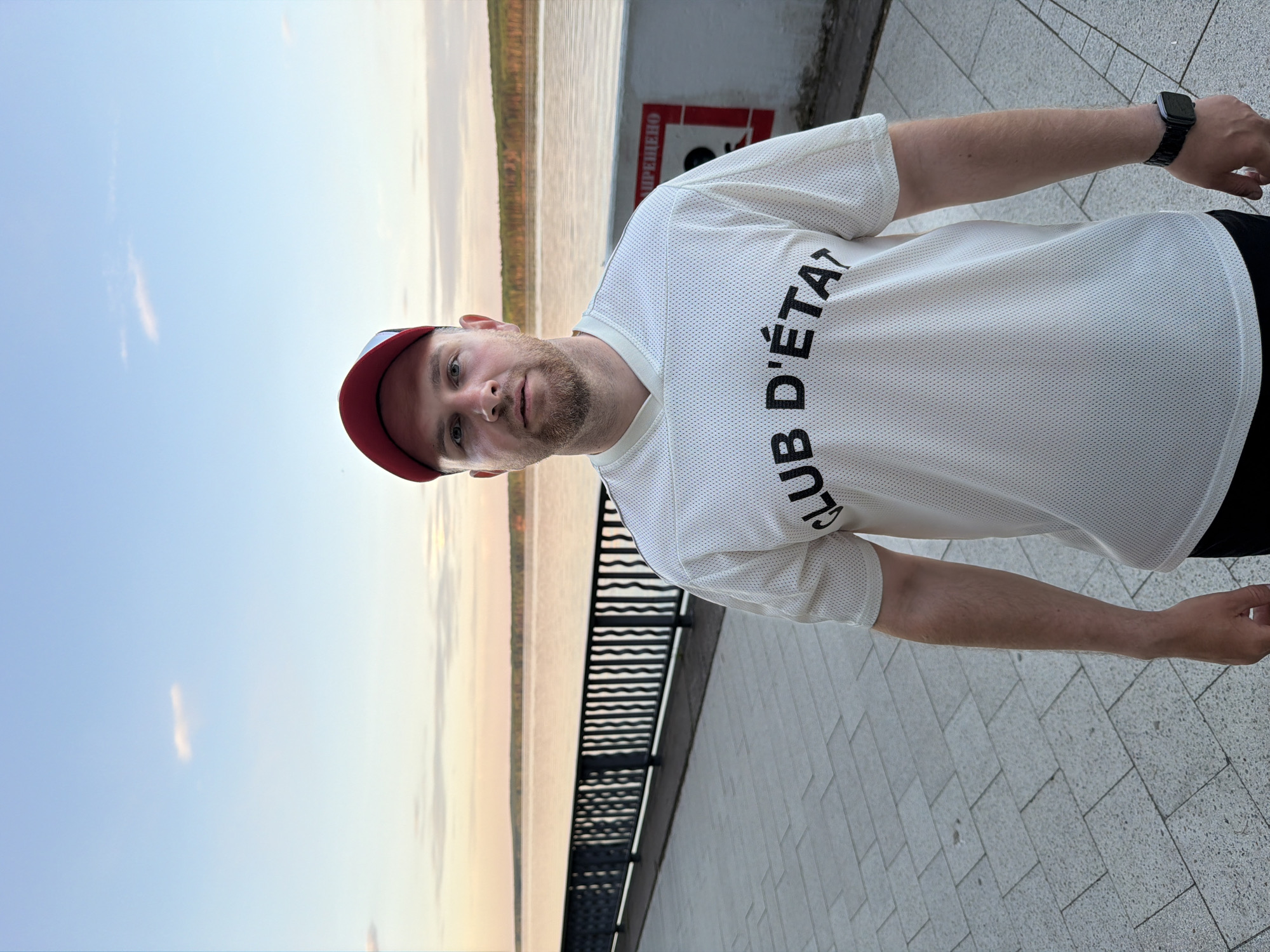 A man stands on a paved surface, likely a sidewalk or promenade, with a metal railing to his left. Behind the railing is a body of water, possibly a river or lake, stretching into the horizon. The sky is light, suggesting either sunrise or sunset. The man is wearing a white, mesh-like t-shirt with the words "CLUB D'ÉTAT" printed on it, a red and blue baseball cap, and a black watch on his left wrist. The location is Chaikovsky, Russia. A sign can be seen on a wall in the background. The man is looking towards the camera.