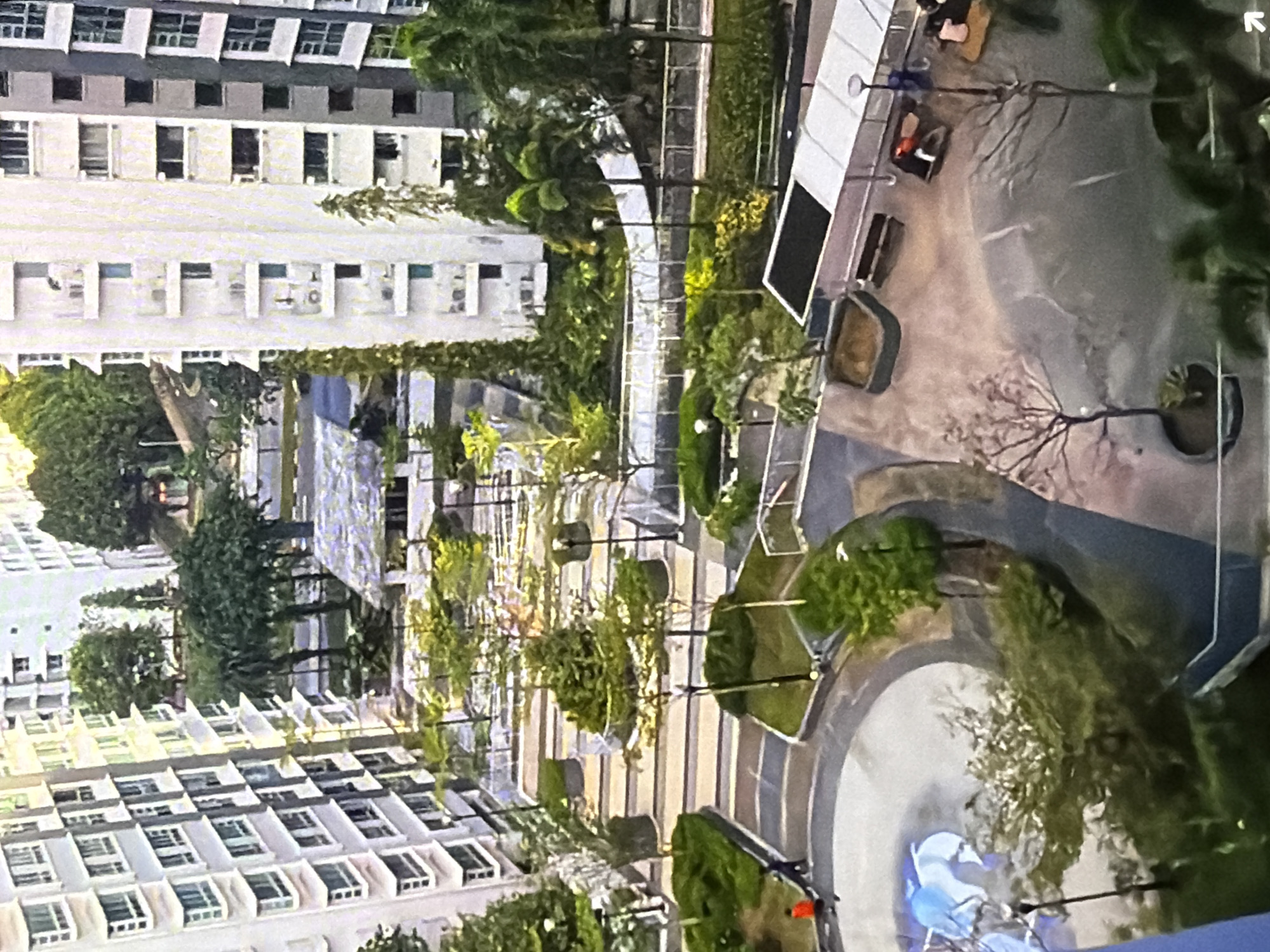 The image displays a high-angle view of an urban courtyard surrounded by multi-story residential buildings. The courtyard features paved pathways, landscaped green spaces with trees and bushes, and several seating areas with benches and covered shelters. A few individuals are visible within the courtyard, one wearing an orange shirt and walking along a pathway. Other people are seated on benches. The architecture of the buildings suggests a modern urban development. The overall scene depicts a well-maintained communal outdoor space within a city.