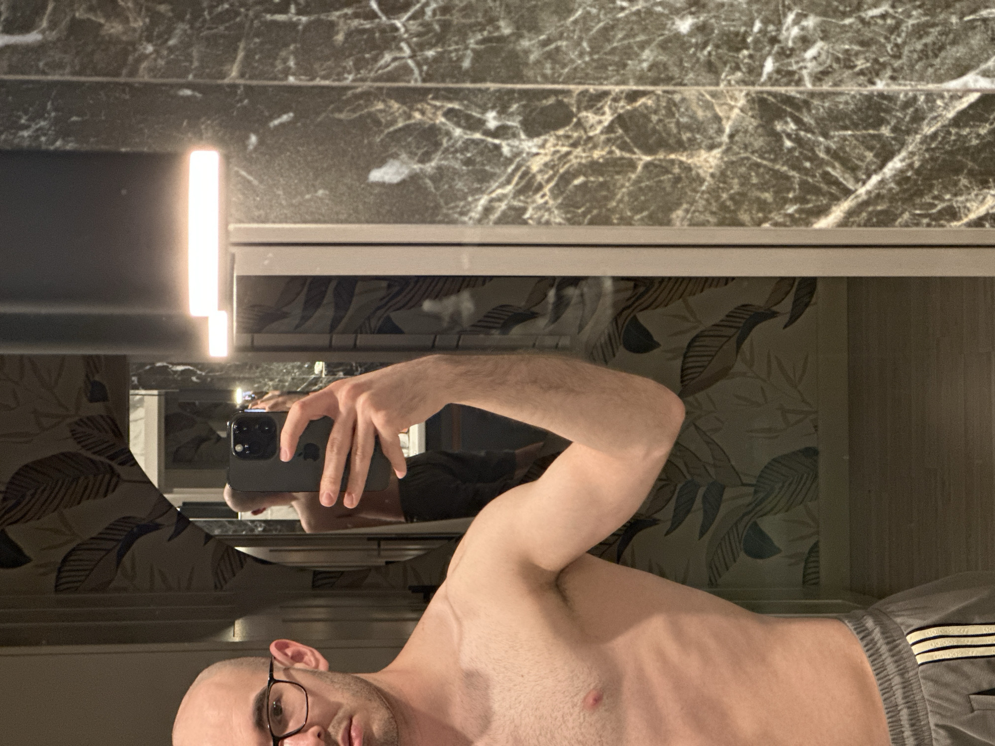 A bald man wearing glasses is taking a selfie in a bathroom mirror. He is shirtless and wearing grey Adidas shorts. The bathroom has marble walls and a vanity with a light on the left side. The mirror reflects a patterned wallpaper with leaves. The location cannot be determined from the image.