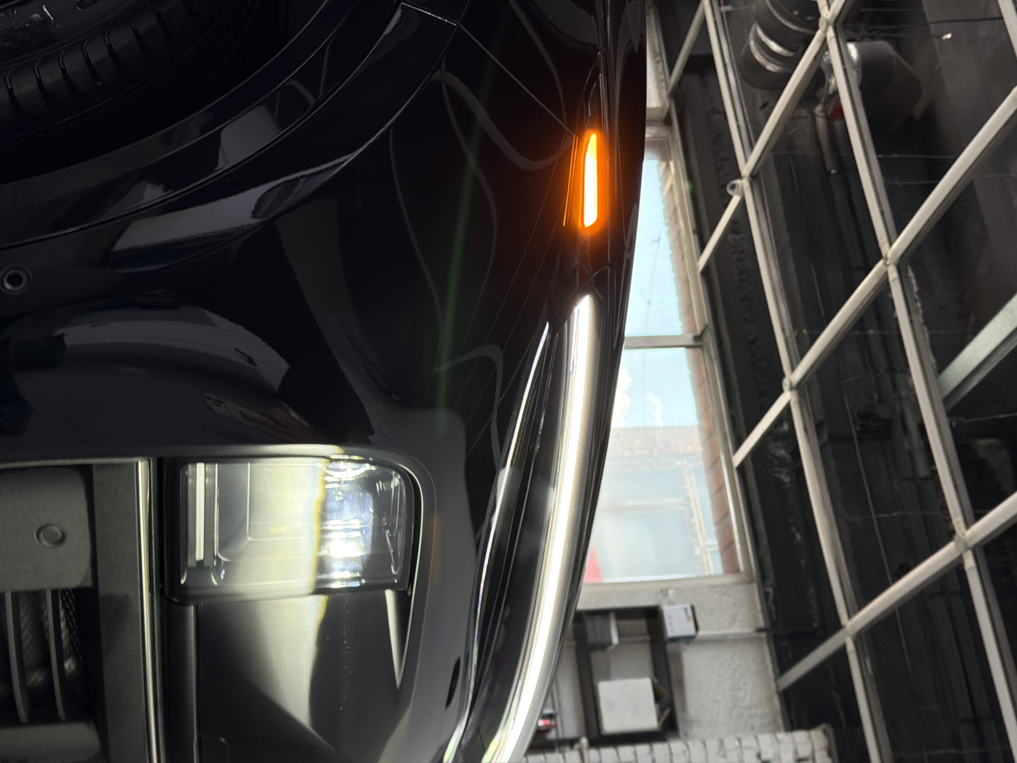 The image depicts a close-up view of the front left side of a black car, likely parked indoors. The car's headlight and a vertical amber indicator light are visible and illuminated. To the right of the car, a window with a grid-like frame is visible, offering a glimpse of the sky and buildings outside. The surrounding environment suggests an industrial or garage setting. There is no indication in the image of the location being in Krasnoyarsk, Russia.