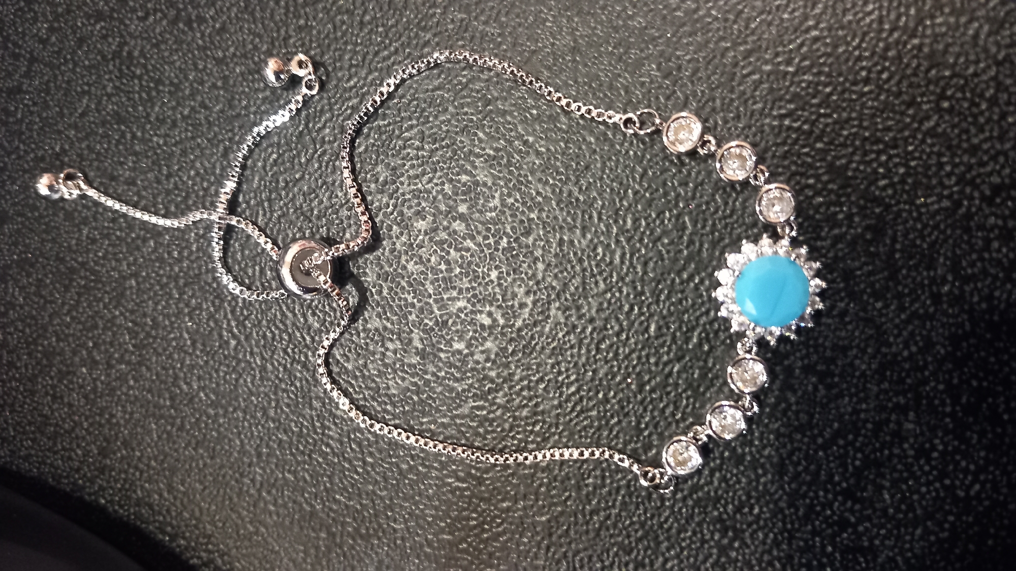 A silver bracelet with a turquoise stone and diamonds is lying on a black textured surface. The bracelet has a delicate chain and a sliding clasp mechanism. The turquoise stone is round and vibrant blue, surrounded by smaller clear stones arranged in a floral pattern. Several other clear stones are also set along the chain. The background is a dark, textured material, possibly faux leather or a similar fabric. There is no indication of the city of Kos, Greece, in the image.