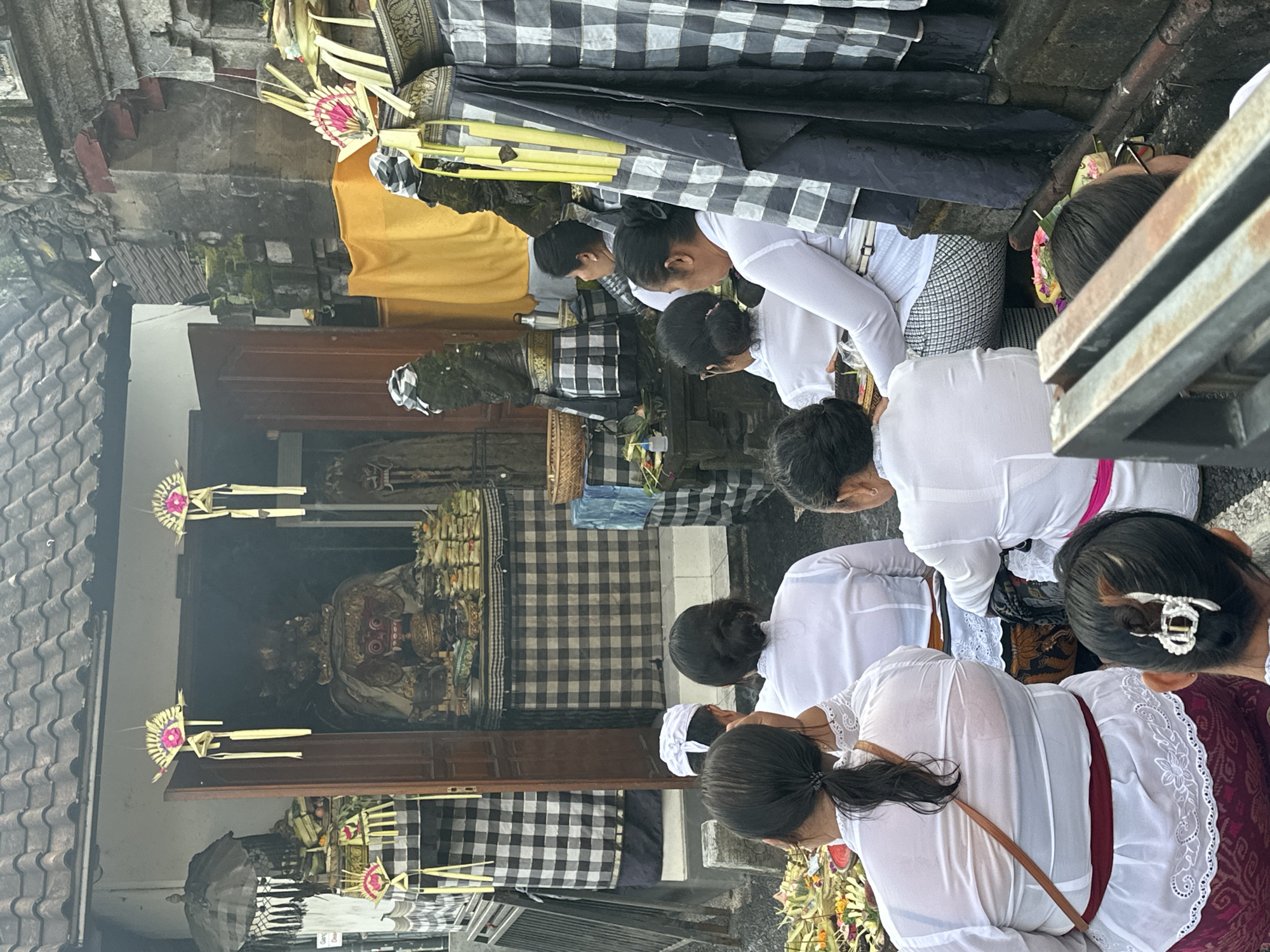 A group of people are gathered in front of an open shrine, possibly a temple or a family altar, in North Kuta, Indonesia. Several individuals are seated, facing the shrine, and appear to be participating in a religious ceremony or offering. They are dressed in traditional white clothing, some with head coverings, and elaborate patterned skirts or sarongs. The shrine itself is adorned with offerings of flowers and woven palm leaf decorations. Inside the shrine, a large, ornate figure with a red face and elaborate headdress is visible. The architecture suggests a Balinese style, with a tiled roof and stone construction. Black and white checkered fabric, a common motif in Balinese culture, is draped around the shrine and on some of the objects. The scene is outdoors, with a slightly overcast sky.